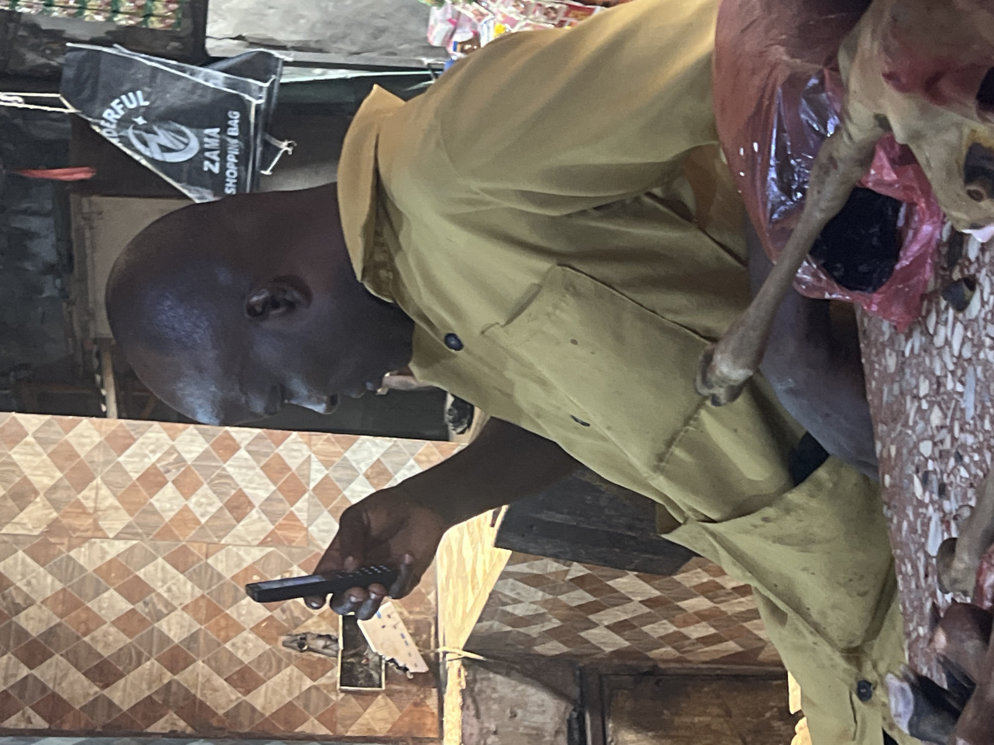 A medium shot shows a dark-skinned man holding what appears to be a remote control. He is wearing a yellow collared shirt. Beside him is a section of what seems to be an animal carcass, partially contained in a red plastic bag. The background includes a tiled wall and a shopping bag with the word "ZAMA" on it. The location is the city of Unknown, Unknown.