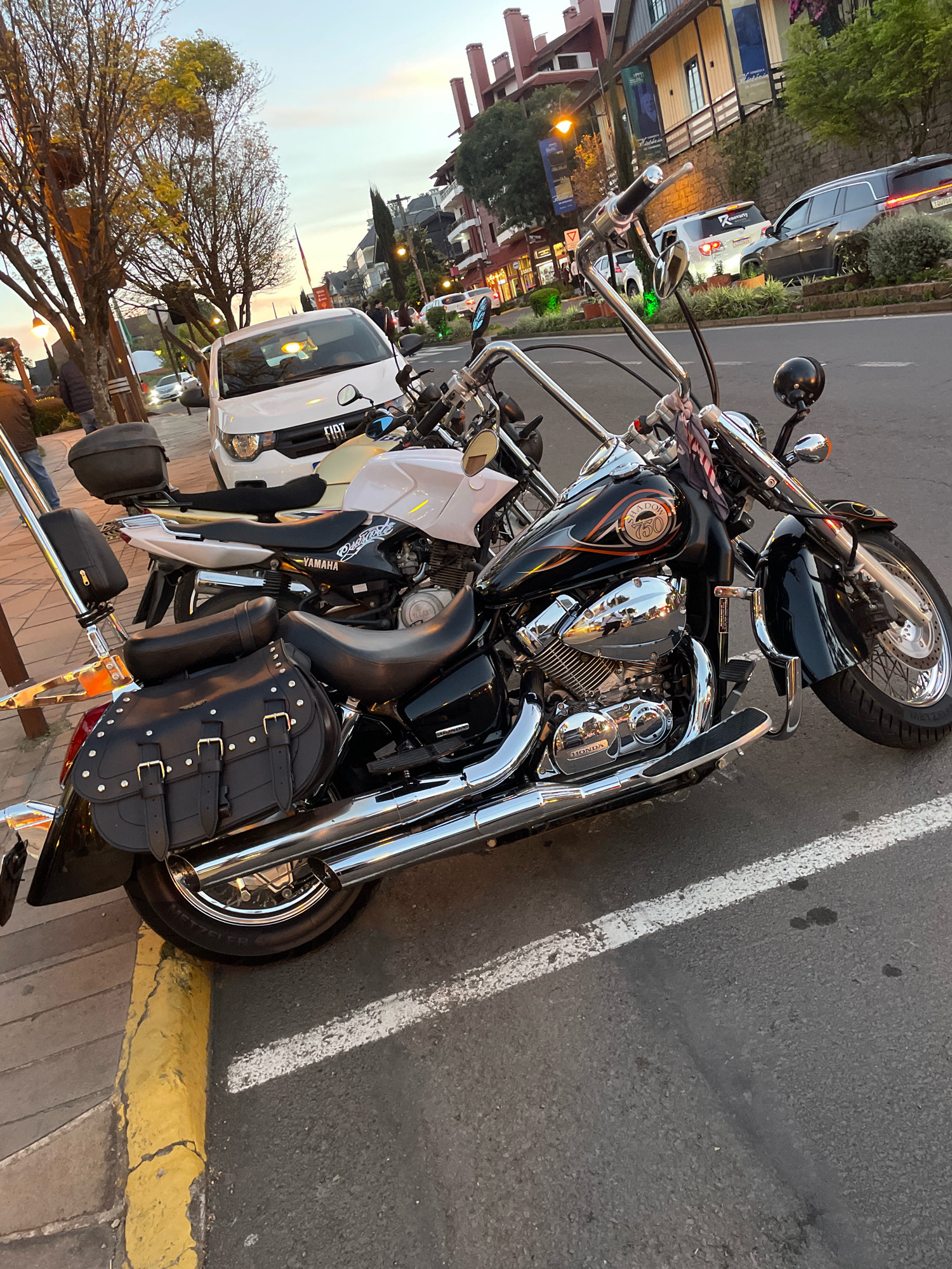 The image depicts a street scene in Gramado, Brazil, likely during the late afternoon or early evening, given the warm lighting and the sky's color. Two motorcycles are prominently featured in the foreground, parked on an asphalt road adjacent to a sidewalk. The motorcycle on the right is a black cruiser with chrome accents, featuring saddlebags and a "Shadow 750" emblem. The motorcycle to its left is a white Yamaha.
Behind the motorcycles, a white Fiat car is parked on the street. The street is lined with buildings, characteristic of a town or city center, with some featuring balconies and awnings. Trees with sparse foliage are visible along the sidewalk, and streetlights are present, indicating a developed urban area. The road has white and yellow markings. The overall impression is of a quiet street with parked vehicles, likely in a commercial or residential district of Gramado. There are no people clearly visible in the immediate foreground, but faint figures can be seen further down the street and on the sidewalk.