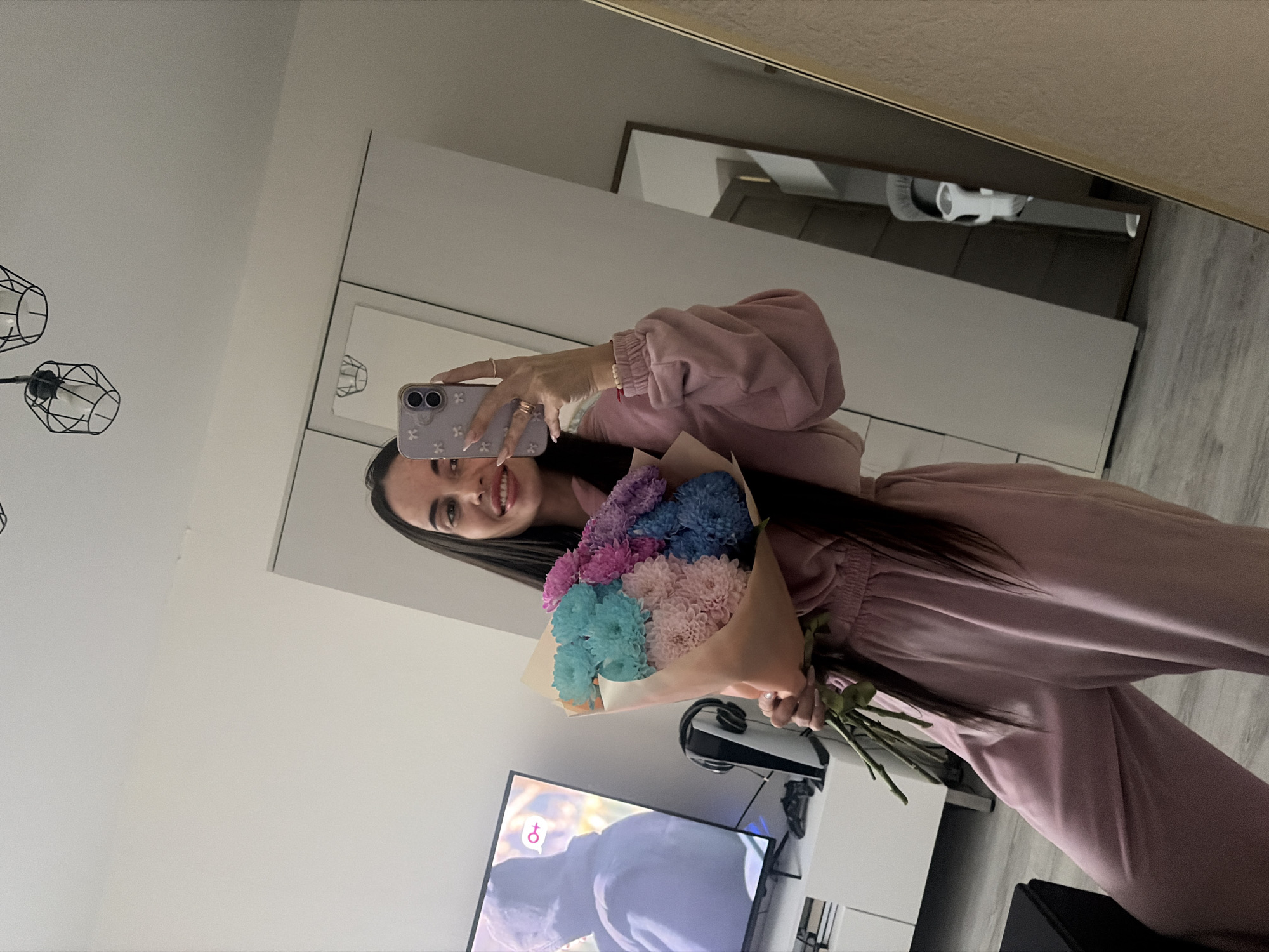 A young woman is taking a selfie in a room. She is wearing a pink sweatsuit and holding a bouquet of colorful flowers. The room appears to be a living room or bedroom, with a television, a white console, and a mirror visible. The television is on, displaying an image. The wall behind her is white. There is a geometric-shaped black lamp on the ceiling. It is not possible to determine the location in Saint Petersburg, Russia from this image.
