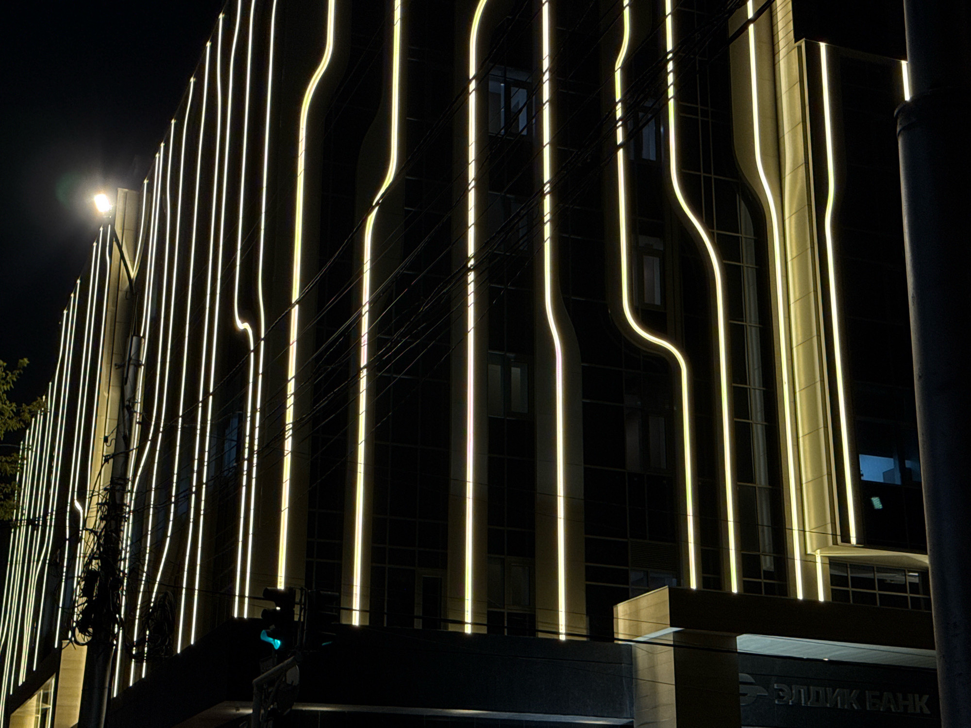 The image shows a modern building at night, likely in Bishkek, Kyrgyzstan, based on the Cyrillic text visible on the building's lower facade which reads "ЭЛДИК БАНК" (Eldik Bank). The building's exterior is adorned with vertical, illuminated neon or LED strips that follow a wavy pattern, creating a striking visual effect against the dark night sky. These lights cast a warm, yellow-white glow. The building features multiple floors with windows that are largely dark, suggesting it is closed or unoccupied at night, though some internal lights might be visible. In the foreground, a traffic light is visible, with its green light illuminated, indicating that traffic is permitted to proceed in at least one direction. There are also electrical wires and poles, typical of an urban streetscape, and a street lamp with a bright, diffused light source to the left of the building. The overall scene is one of urban nighttime architecture, illuminated by artificial light.