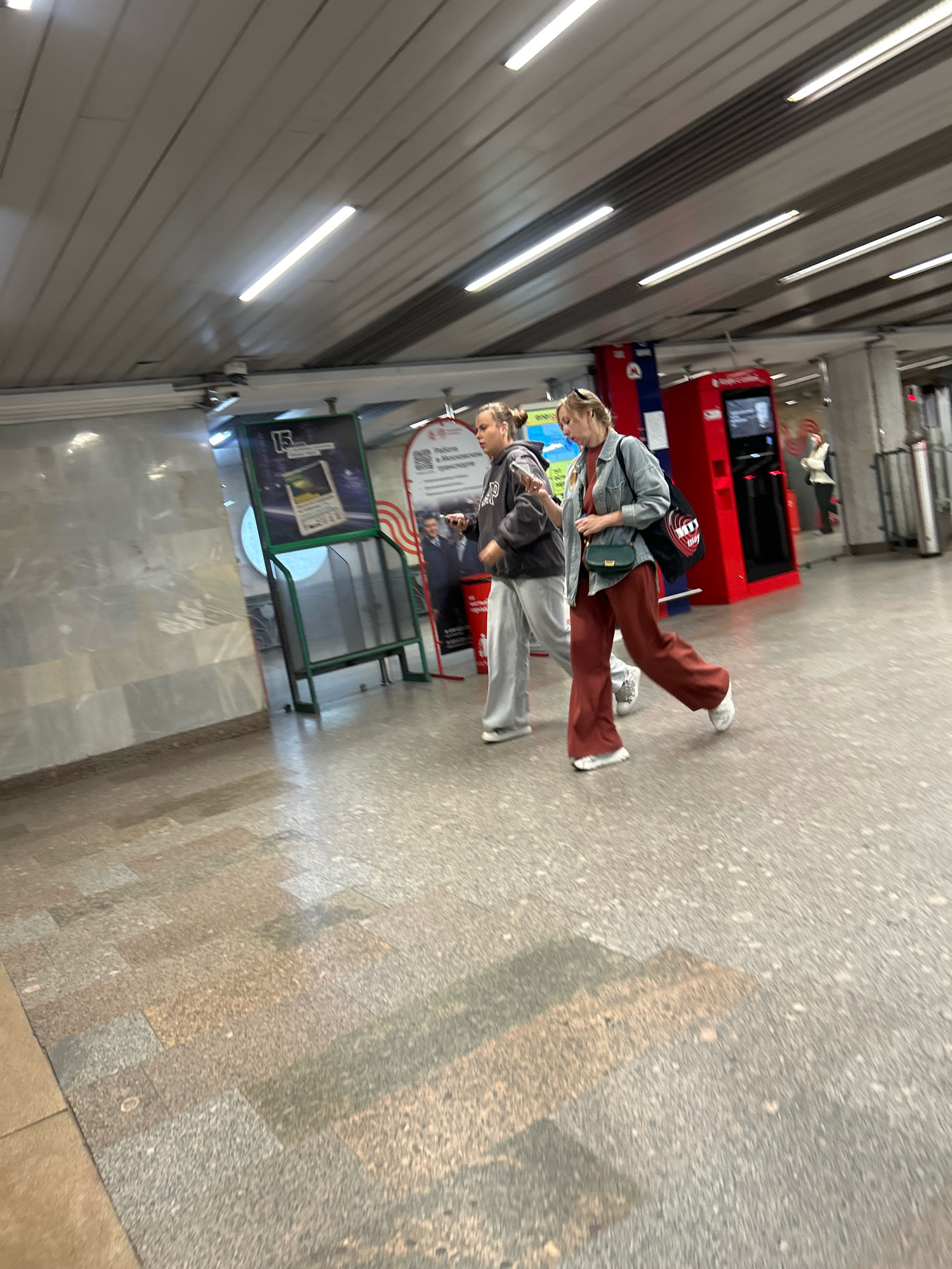 Two young women walk side-by-side through a brightly lit, modern subway station in Moscow, Russia. The woman on the left wears a gray hoodie and light gray sweatpants, with her hair pulled back in a bun. She is looking down at her phone. The woman on the right, with blonde hair pulled back, wears a denim jacket over an orange t-shirt and rust-colored wide-leg pants. She carries a black tote bag over her shoulder and a small green purse. Both women are walking on a textured, speckled floor, with large, light-colored marble-like tiles on the left wall. Behind them, a bright red vending machine stands next to a green display rack holding a poster. Fluorescent lights line the angled ceiling, and a silver ventilation system is visible. The overall impression is one of urban transit and everyday life.