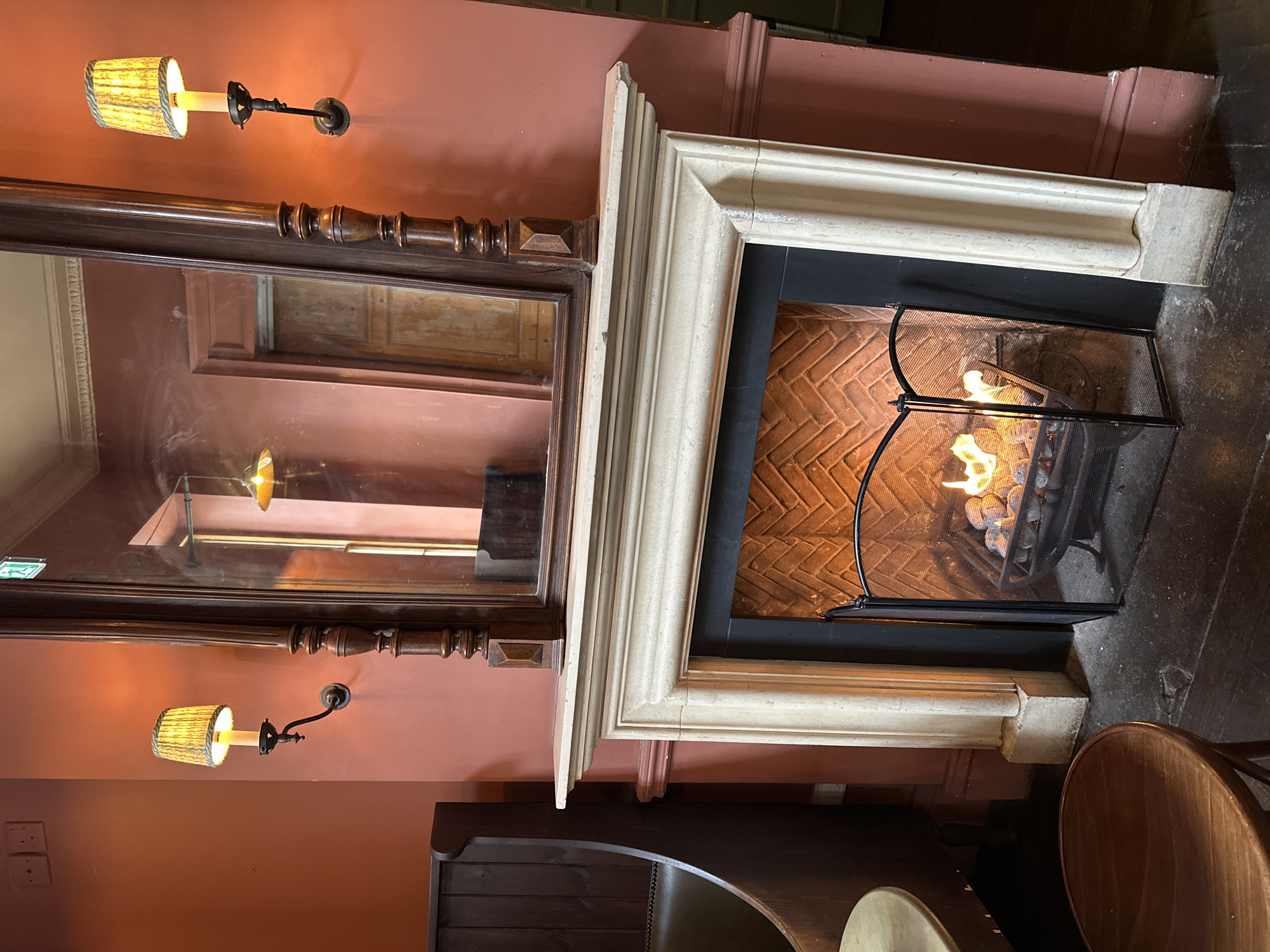 The image shows a fireplace with a white mantle and a fire burning inside. A dark wood-framed mirror hangs above the fireplace. Sconces with small, patterned shades flank the mirror on a salmon-colored wall. Below, a dark wood sideboard is visible. The fireplace is in a room with other dark wood furnishings including a round table and a chair.
Without additional information, pinpointing a specific location in London is not possible.