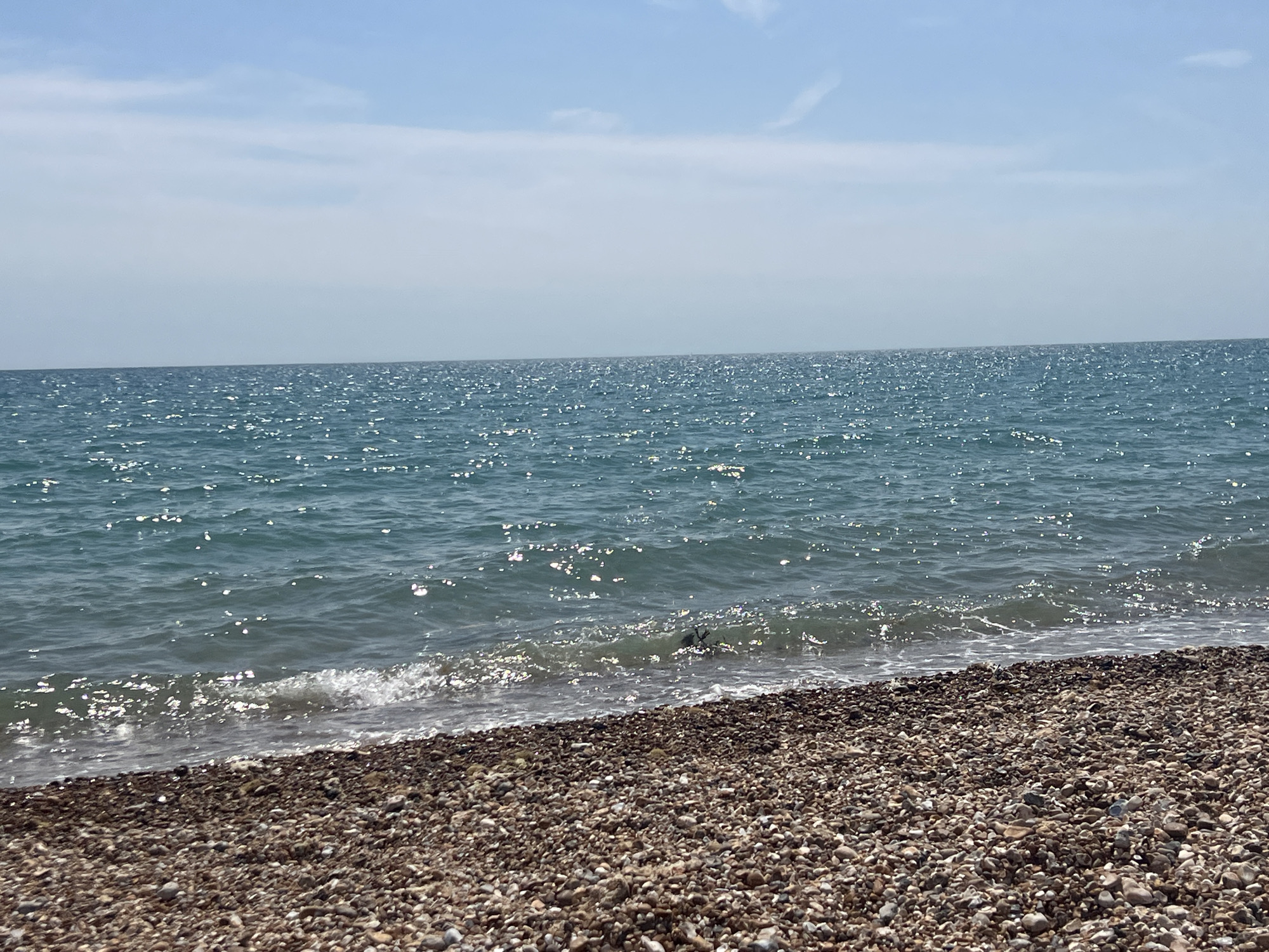 The image shows a pebble beach with the sea in the background. The sea is a deep blue-green and reflects the sunlight, creating a shimmering effect. There are small waves lapping at the shore. The beach is covered in a variety of small, smooth pebbles in shades of brown, grey, and white. The sky is a pale blue with a few wispy clouds. The scene appears to be a calm and sunny day at the seaside. Based on the presence of a pebble beach, the location is likely to be a coastal area. Given the details, it is plausible this image was taken in Arun, United Kingdom, which is known for its coastline with pebble beaches.