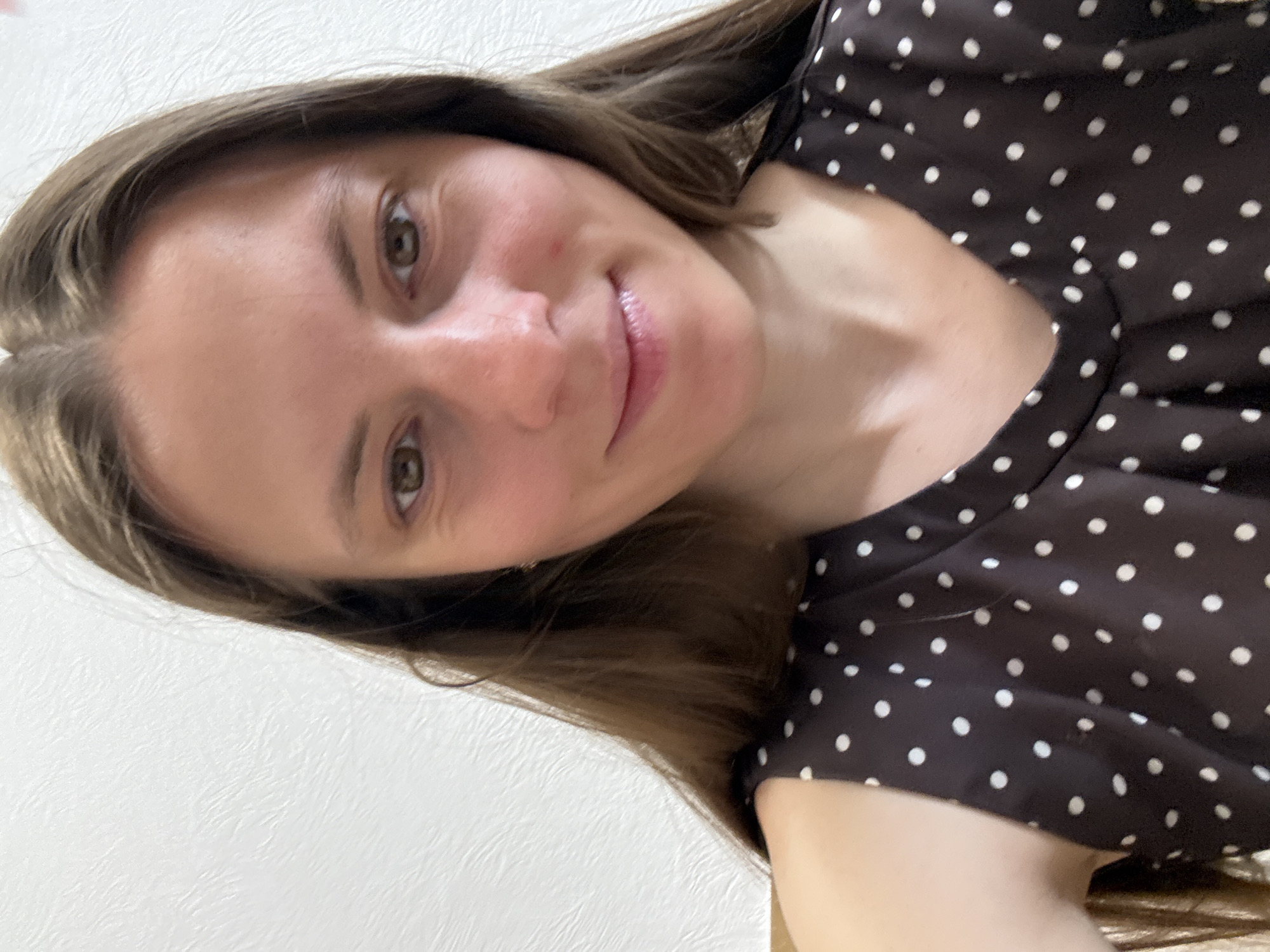 A woman with long brown hair is shown in a selfie, wearing a brown polka dot dress. The background is a plain white wall. There is no indication of the location being Chaikovsky, Russia.