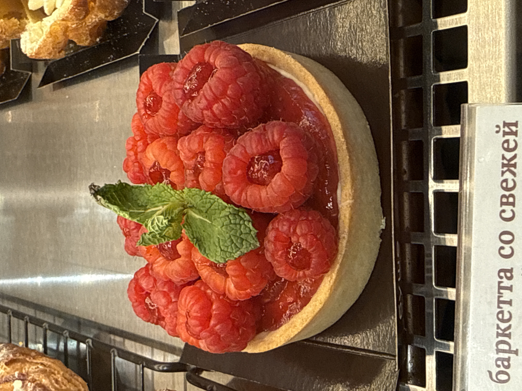 The image shows a raspberry tart with a mint sprig garnish displayed in a bakery or pastry shop. The tart has a light-colored crust filled with a layer of red filling, topped with fresh raspberries. A small mint sprig is positioned on top. The tart is on a dark metal surface, likely a display tray within a refrigerated case. A sign in Russian is visible on the right, partially obscured, likely indicating the name and possibly the ingredients of the tart ("баркетта со свежей" suggests "tart with fresh"). The location is suggested to be in Saint Petersburg, Russia. Other baked goods are visible in the background.