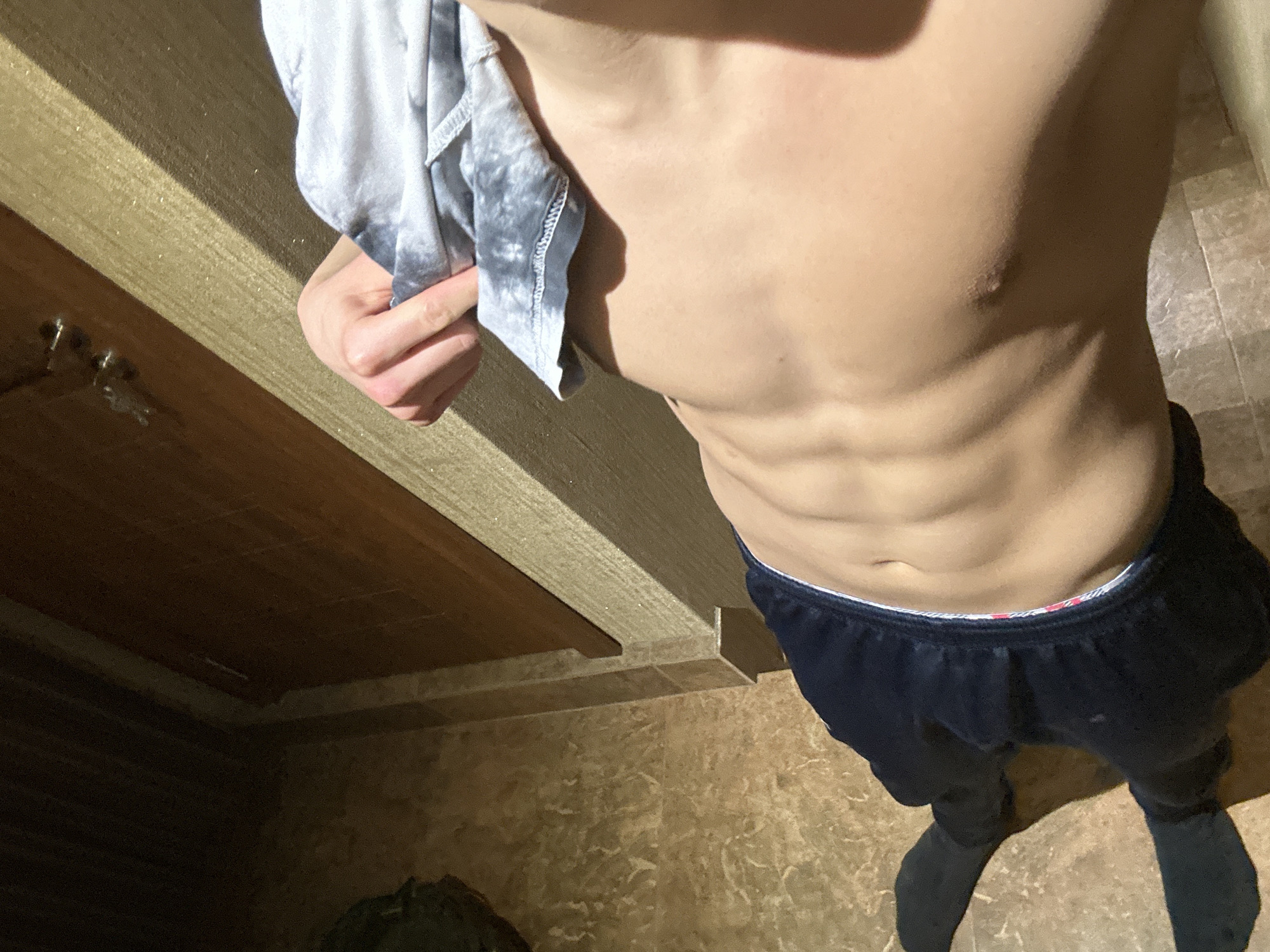 A person is standing in what appears to be a bathroom. They are wearing dark-colored sweatpants and no shirt, revealing their torso. They are holding a gray and white patterned garment. The bathroom has a wooden vanity countertop and patterned floor tiles. No information indicates the location is in Burgas, Bulgaria.