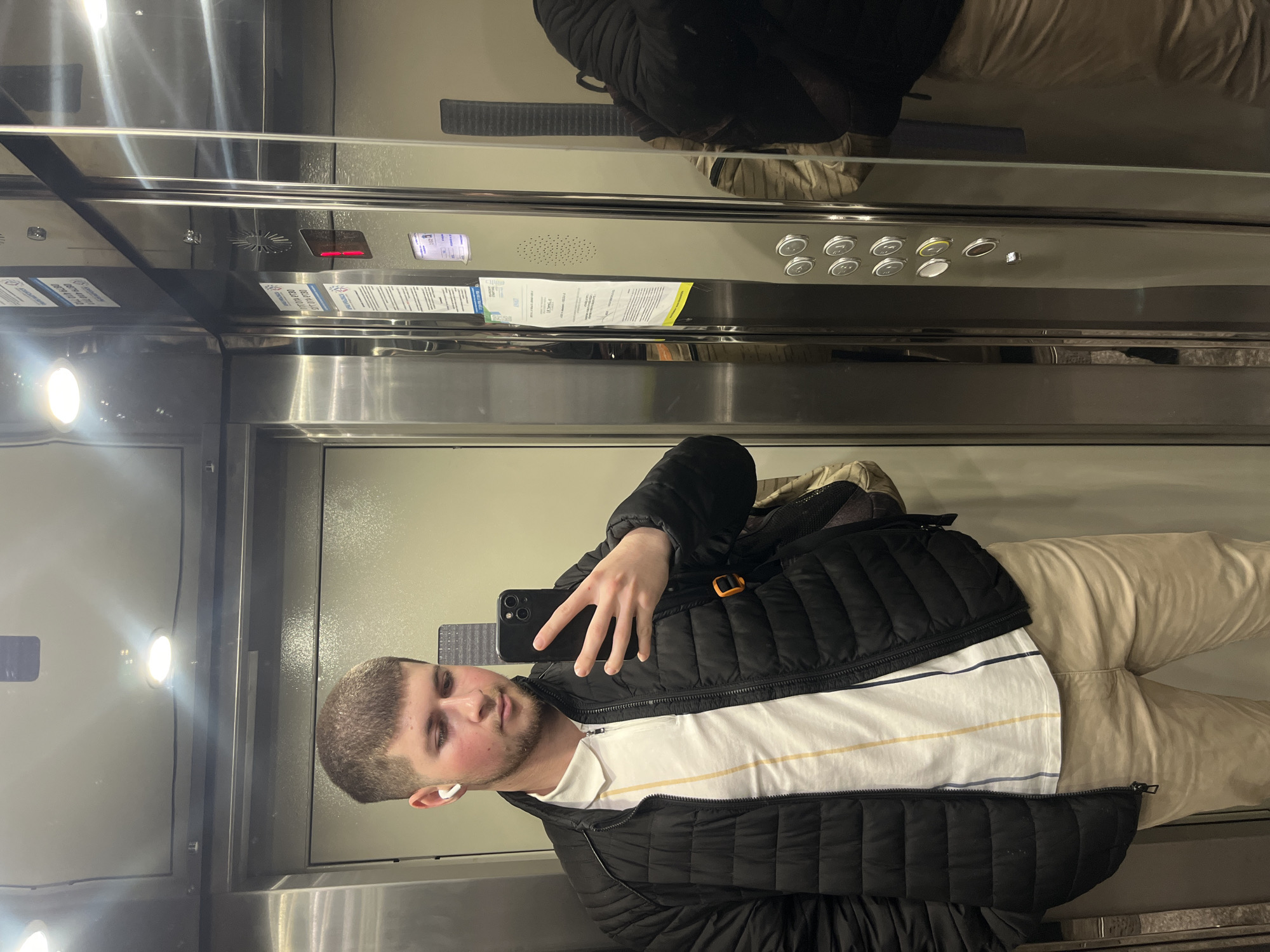 A male is taking a selfie in an elevator. The elevator has a silver interior with buttons and a screen. The man is wearing a black jacket, a striped white shirt, and beige pants. He has an earbud in his left ear. The exact location in Varna, Bulgaria cannot be determined from this image.
