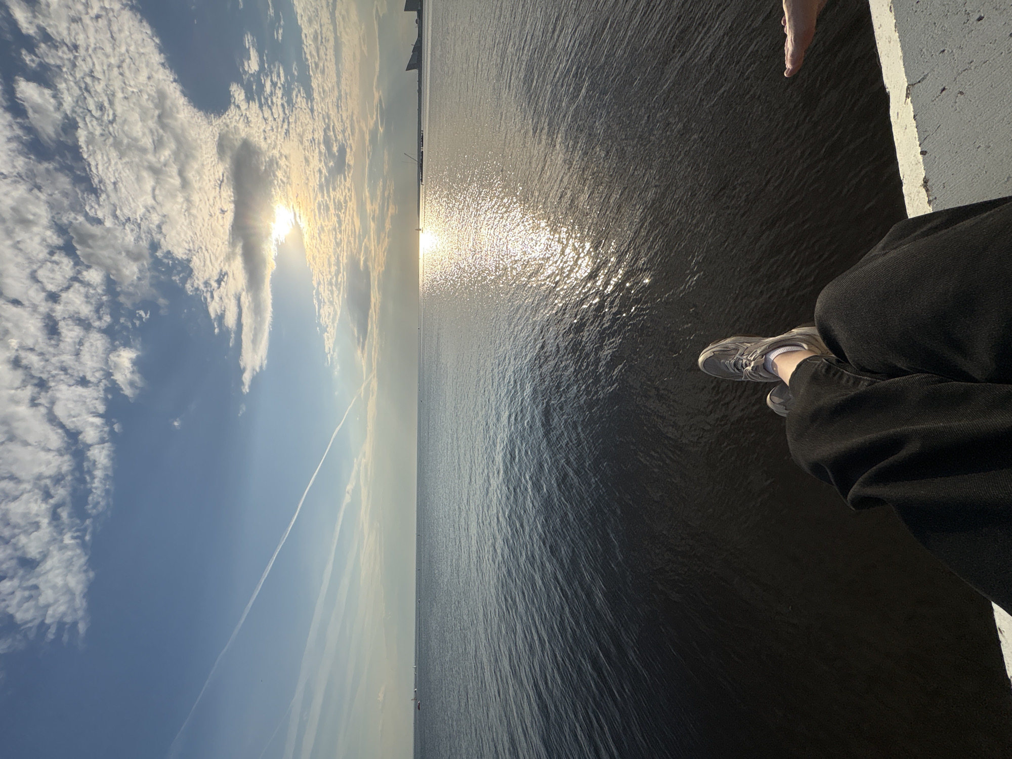 This image captures a serene waterscape at either sunrise or sunset, viewed from a slightly elevated perspective. The scene is bisected horizontally, with the sky on the left and the expansive body of water on the right.
The sky on the left transitions from a clear, light blue at the bottom to a more dynamic canvas towards the top, where the sun is low on the horizon. The sun, partially obscured by bright, scattered cumulus clouds, casts brilliant rays and illuminates the surrounding atmosphere. A distinct, long contrail from an aircraft stretches across the clearer blue sky, adding a linear element to the vastness.
The large body of water, likely a sea or major river such as the Gulf of Finland or Neva River in Saint Petersburg, Russia, dominates the right portion of the image. Its surface is a deep, dark blue-grey, reflecting the soft light of the sky but also showing intense sparkling reflections from the low-lying sun. The sun's reflection creates a radiant path across the water, drawing the eye towards the horizon where faint outlines of distant land or structures, possibly boats or a city skyline, are visible.
In the bottom right corner of the frame, a person is seated on a white concrete ledge or barrier. Their legs are seen dangling, with one leg clad in dark pants and a grey sneaker with white socks. A bare foot is partially visible above it, suggesting either the other foot of the same person or a second person also seated on the ledge. The activity appears to be peaceful observation of the natural surroundings.
The time of day is clearly indicated by the low sun, suggesting either an early morning or late evening scene, most likely sunset given the warm hues. The weather is fair with scattered clouds. No text is visible in the image. The overall mood is one of tranquility and contemplation, characteristic of observing a scenic vista.