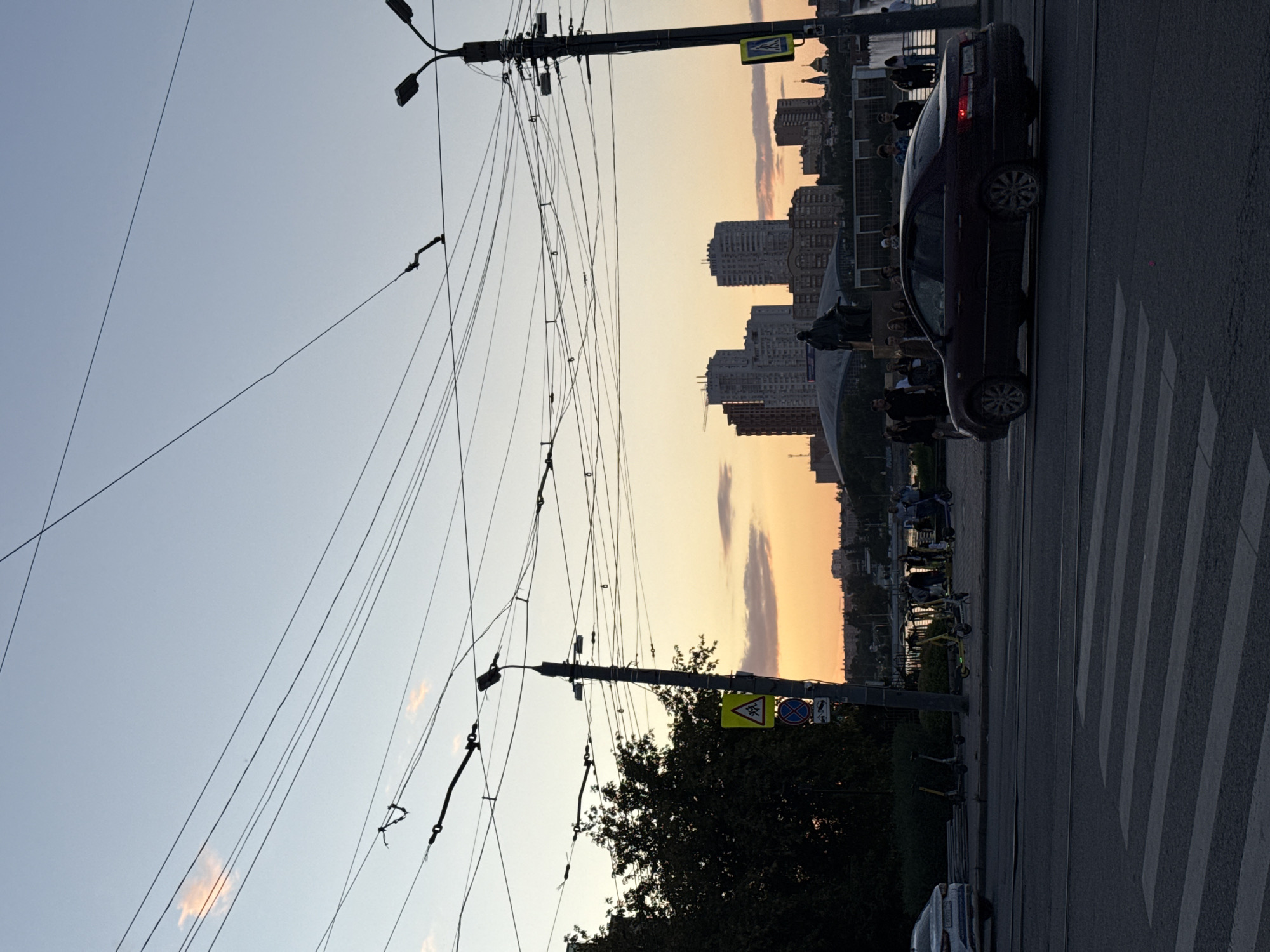 This image captures a street scene in Chelyabinsk, Russia, during the twilight hours. A maroon sedan is seen driving across a pedestrian crossing on a paved road. In the background, several people are visible near a body of water, with various buildings and a statue in the distance. The sky displays a gradient of colors, transitioning from light blue at the top to orange and pink hues near the horizon, indicative of sunset. Overhead, a complex network of power lines crisscrosses the frame. On the left side of the image, a tree and a street sign are present. The overall atmosphere suggests a typical urban evening with traffic and pedestrian activity.