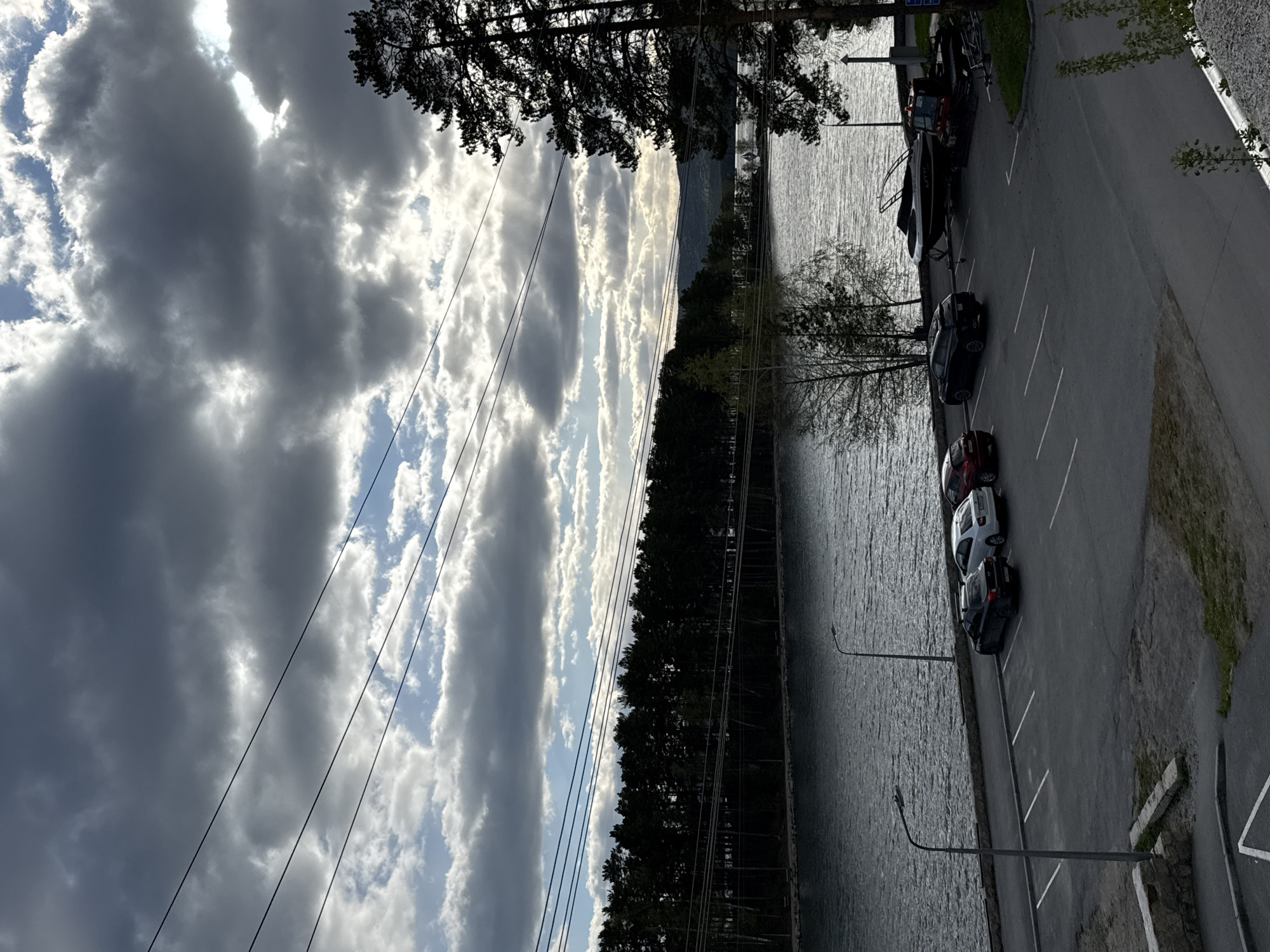 The image captures a view of a parking area adjacent to a body of water in Dom Otdykha Zolotoy Plyazh, Russia. Several cars are parked along a paved road, and a boat is also parked. A line of trees separates the parking area from the water. Power lines are visible in the sky, which is filled with clouds. The sun is shining through some areas of the clouds.