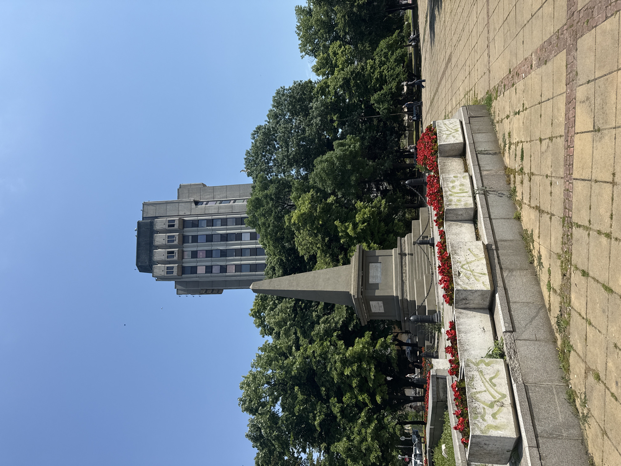 The image, captured with a vertical orientation (possibly a landscape shot rotated 90 degrees counter-clockwise), depicts a sunny urban scene featuring a monument, lush trees, a tall building, and paved walkways.
In the foreground (right side of the image as presented), a broad paved area consists of light-colored square tiles interspersed with reddish brickwork. Weeds grow visibly between some of the tiles. A series of tiered concrete planters runs parallel to the pavement, filled with vibrant red flowers, likely begonias. Several of these planters show light-colored, abstract graffiti or markings. Black bollards connected by chains are also visible near the planters.
Towards the center of the image stands a prominent grey stone obelisk-style monument with a square base. Plaques are affixed to its sides, though the text is unreadable. Surrounding the monument and extending across the middle section of the image are numerous dense, green deciduous trees, suggesting a park or wooded area. Several people are faintly visible among the trees, either sitting or walking, indicating a casual public space.
In the background (left side of the image as presented), a tall, modern-looking grey building rises against a clear, bright blue sky. Its multi-story facade features many windows. The building partially extends out of the frame at the top. The bright sunlight creates clear shadows, emphasizing the sunny weather. A small bird is visible flying in the expansive blue sky.