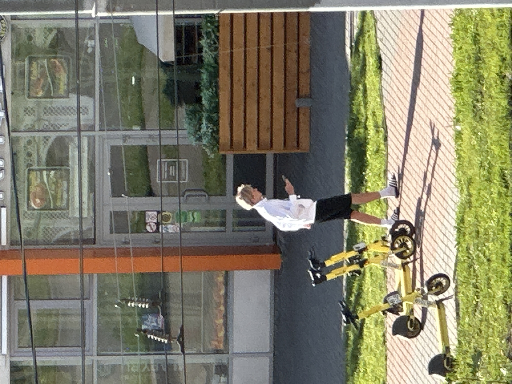 A blonde male wearing a white long-sleeved shirt and black shorts walks on a sidewalk, looking at his phone. He is wearing white socks with black stripes and black athletic shoes. In the foreground, three yellow electric scooters are parked on the grass and sidewalk. The scooters have black handlebars and wheels. Behind the scooters and the man is a building with a large glass storefront. The building has an orange pillar on the left side. In front of the building, there is a long wooden planter box filled with green foliage. The image appears to have been taken during the daytime, with shadows cast by the man and the scooters on the sidewalk. The location is identified as Podolsk, Russia.