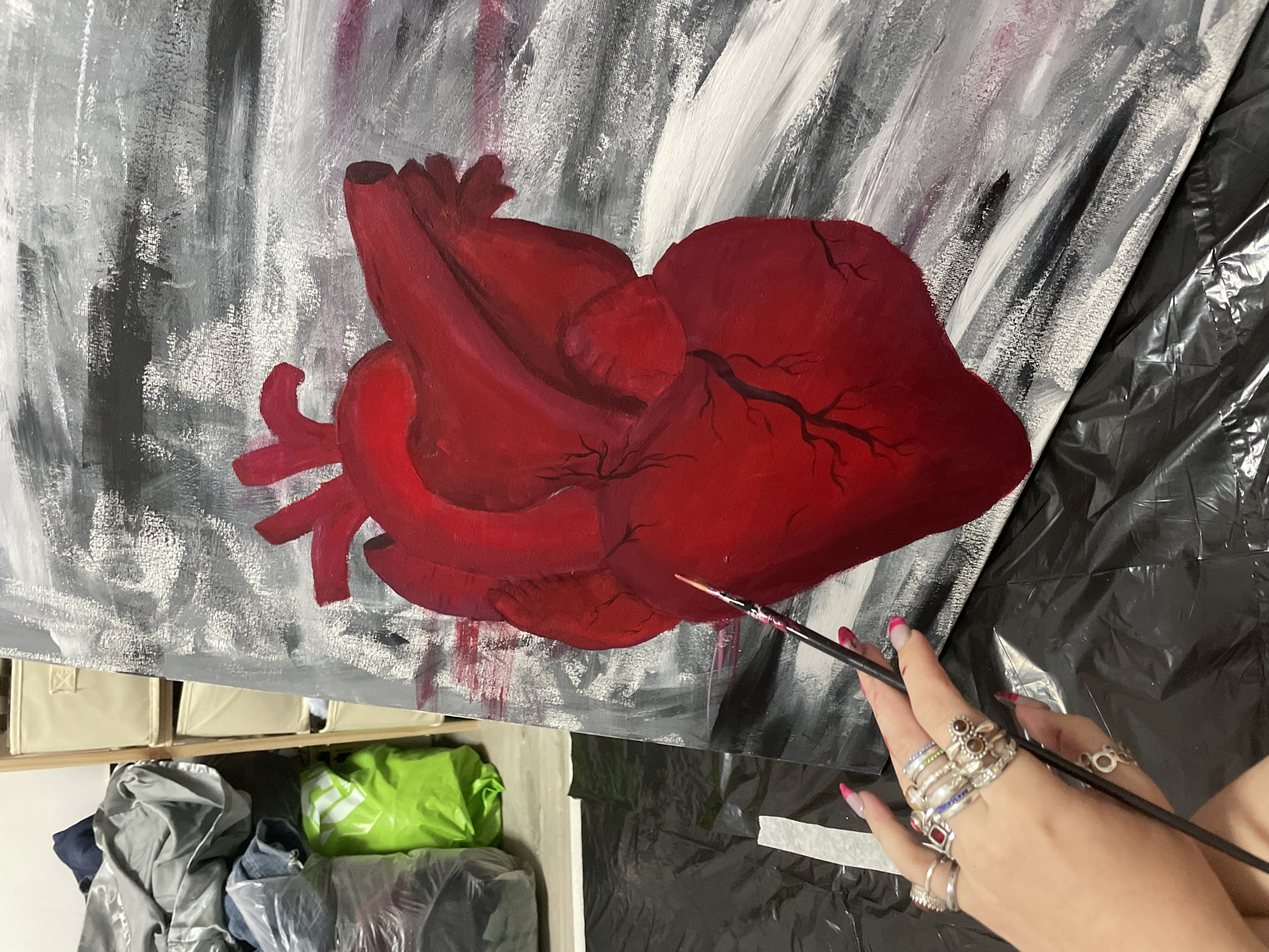The image shows a partially completed painting of a human heart in shades of red, set against a textured background of white, gray, and some purple. The heart has visible veins branching out on its surface. A person is holding a paintbrush in their right hand, which is adorned with multiple rings. The painting is on a canvas, likely resting on a surface covered with a black plastic sheet. Below the canvas, there are plastic storage bins, some filled with clothing or fabric, and a folded green bag. The location in Raanana, Israel is not discernible from the image.