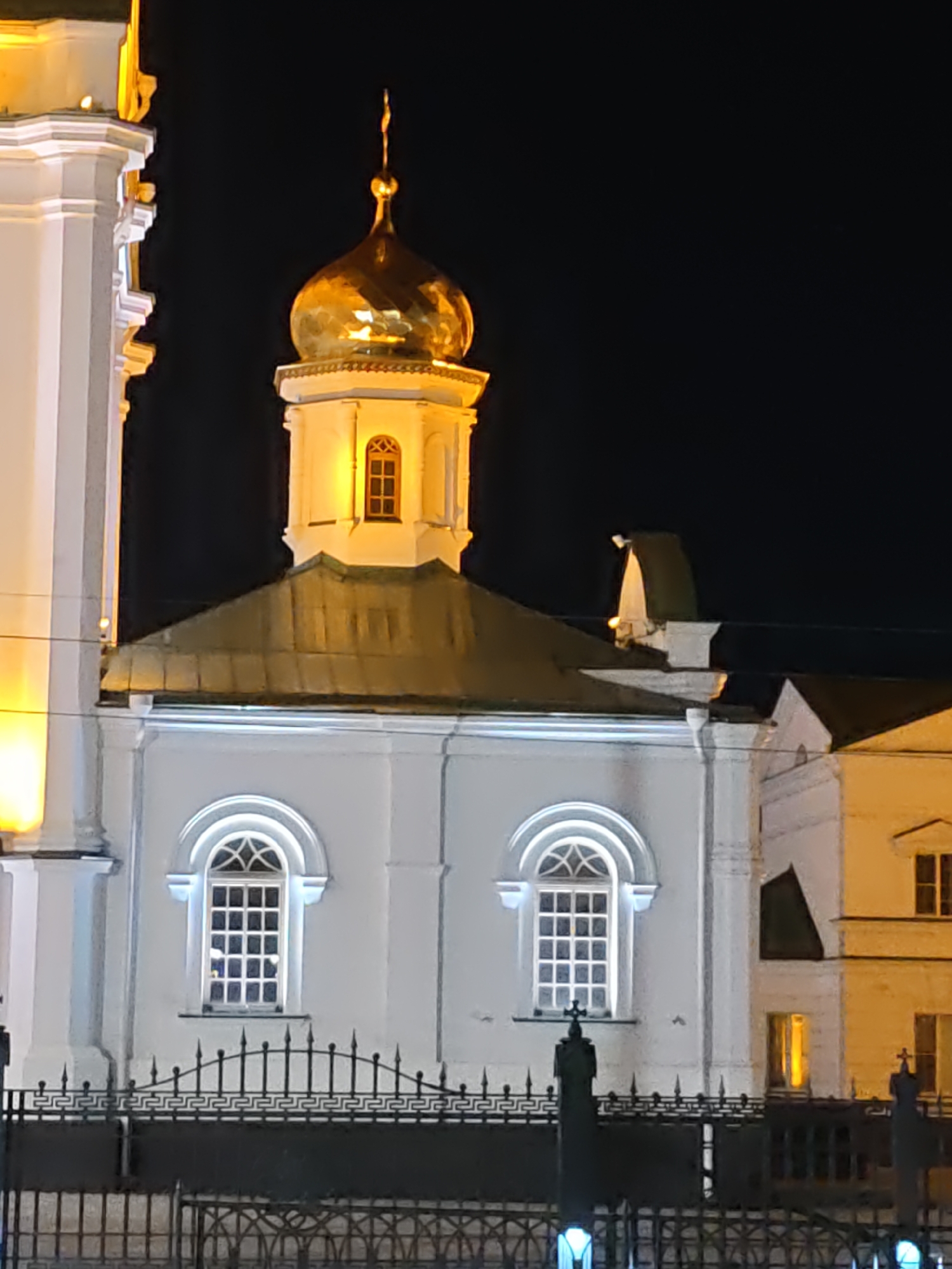 A close-up, low-angle, night-time photograph shows the exterior of a building, likely a church, with distinct architectural features. The structure is predominantly white and illuminated by artificial lights, highlighting its facade.
Dominating the upper center of the image is a prominent golden dome, topped with a spire. The dome appears to be faceted and reflects the light. Below the dome, a section of the building is visible, featuring a square-shaped window with a cross-like design within its panes, and this section is also brightly lit.
The main body of the building, visible in the lower half of the frame, is also white. It features two large, arched windows. Each window is divided into a grid of many small squares, suggesting a stained-glass or decorative design, and they are illuminated from within. The windows are flanked by white pilasters or decorative elements.
A dark, ornate metal fence runs across the bottom of the image, in front of the building. The fence has a repeating geometric pattern and is topped with pointed finials. Beyond the main white structure, other parts of the building complex are visible, including a portion of another white building to the right, with at least one lit window, and hints of further illuminated structures in the background. The sky is completely dark, indicating it is night. The overall impression is of a grand, well-lit religious or historical building.
The location is identified as Rostov-on-Don, Russia, and the specific structure is likely the Assumption Cathedral, a prominent landmark in the city.