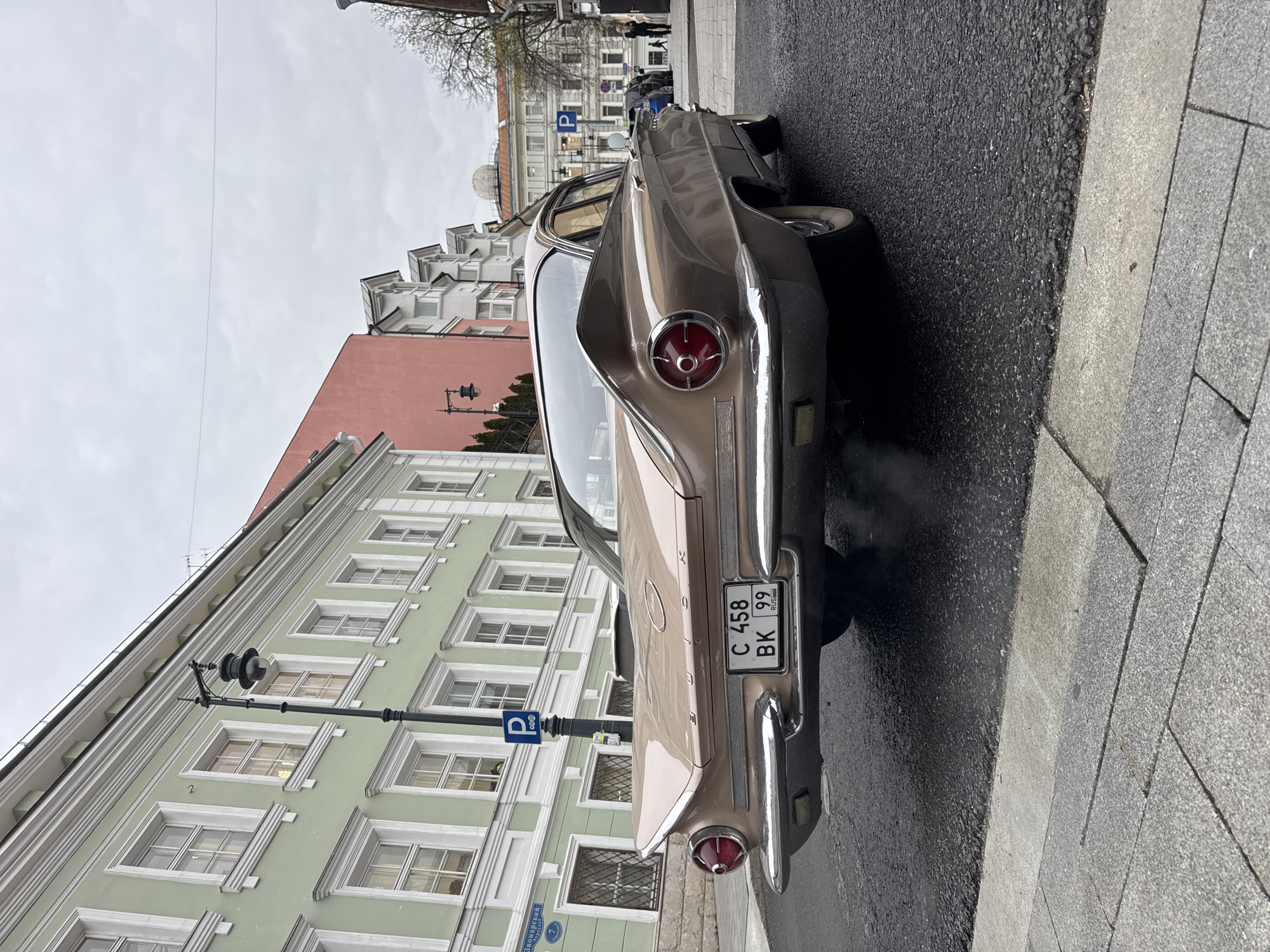 The image shows the rear of a vintage, light brown car parked on a wet street in Moscow, Russia. The license plate reads "C 458 BK 99 RUS". The street is paved with asphalt, and a grey stone sidewalk is visible on the right. A light-green building with white trim and multiple windows stands adjacent to the car. A black lamp post with a parking sign is positioned near the building. Across the street, there is a peach-colored building. Pedestrians can be seen further down the street. The sky is overcast. The road sign visible is Заполярный проезд.