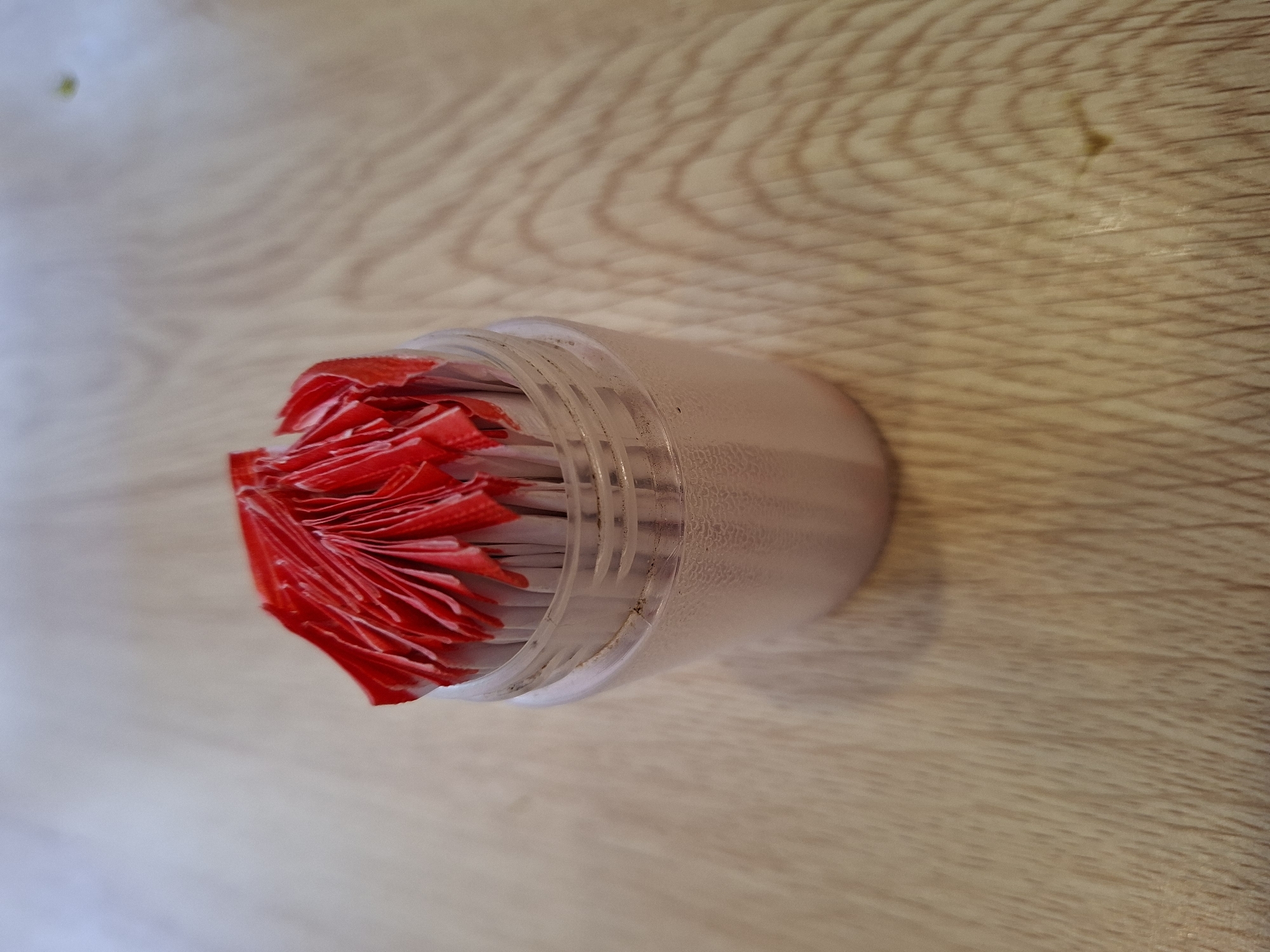 A close-up, eye-level shot shows a clear plastic container filled with toothpicks, their tips wrapped in crinkled red paper. The container is situated on a light-colored wooden surface, possibly a table, with visible wood grain and subtle shadows cast from the object and an unseen light source. The background is a blurred, neutral tone. The image does not provide any information or visual cues that can be used to determine its location within Banjaratma, Indonesia.