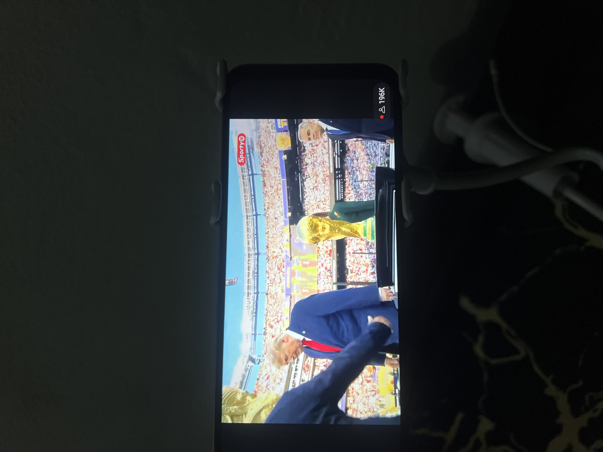 The image shows a smartphone screen displaying a broadcast of a sporting event. On the screen, two men are visible on a stage in front of a large crowd in a stadium. The man on the left appears to be Donald Trump, wearing a blue suit and red tie, and the man on the right is wearing a dark suit and blue tie. A golden trophy, likely the FIFA World Cup, is prominently displayed on the stage. The background shows spectators filling the stadium stands. Overlaid on the screen are broadcast graphics, including a "Sporty Live" logo in the upper right corner and a viewer count of "196K" with an icon of two people in the lower right corner. The smartphone itself is held by a dark-colored stand. The scene is dimly lit, suggesting it is evening or indoors. There is no indication of the location being the city of Jos, Nigeria.