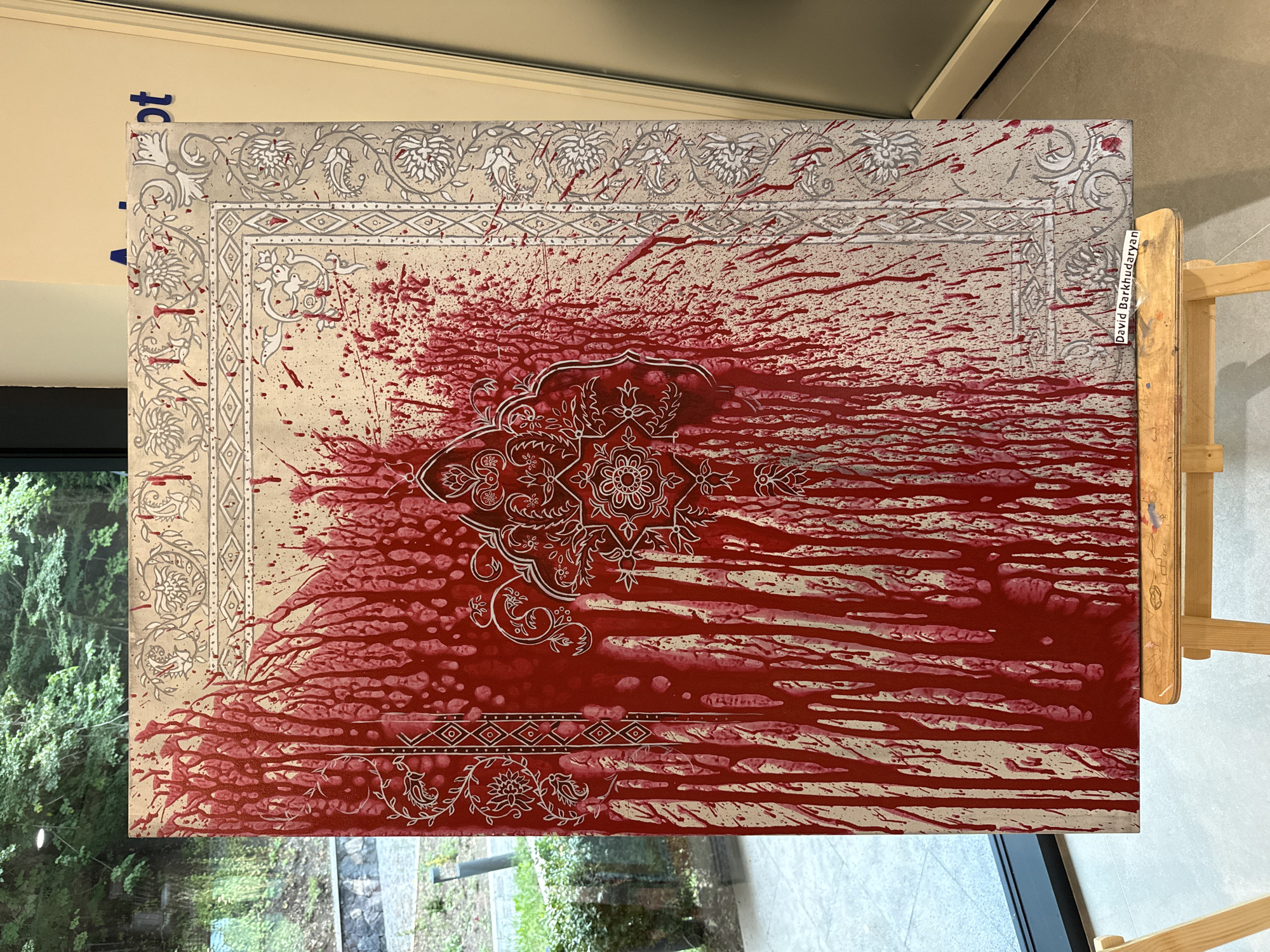 The photograph captures a close-up view of a painting on an easel. The painting is rectangular, oriented vertically, and displays a distressed appearance with numerous red drippings and splatters across a light-colored background. Intricate white patterns, resembling floral motifs and geometric borders, are visible beneath the red markings. A signature, "David Barkhudaryan," is affixed to the bottom edge of the painting. The painting is displayed indoors, likely in an art gallery or exhibition space, with a blurred background that suggests windows looking out onto greenery and a street, possibly in Dilijan, Armenia, a city known for its natural beauty and artistic community. The lighting is even, highlighting the textures and details of the artwork.