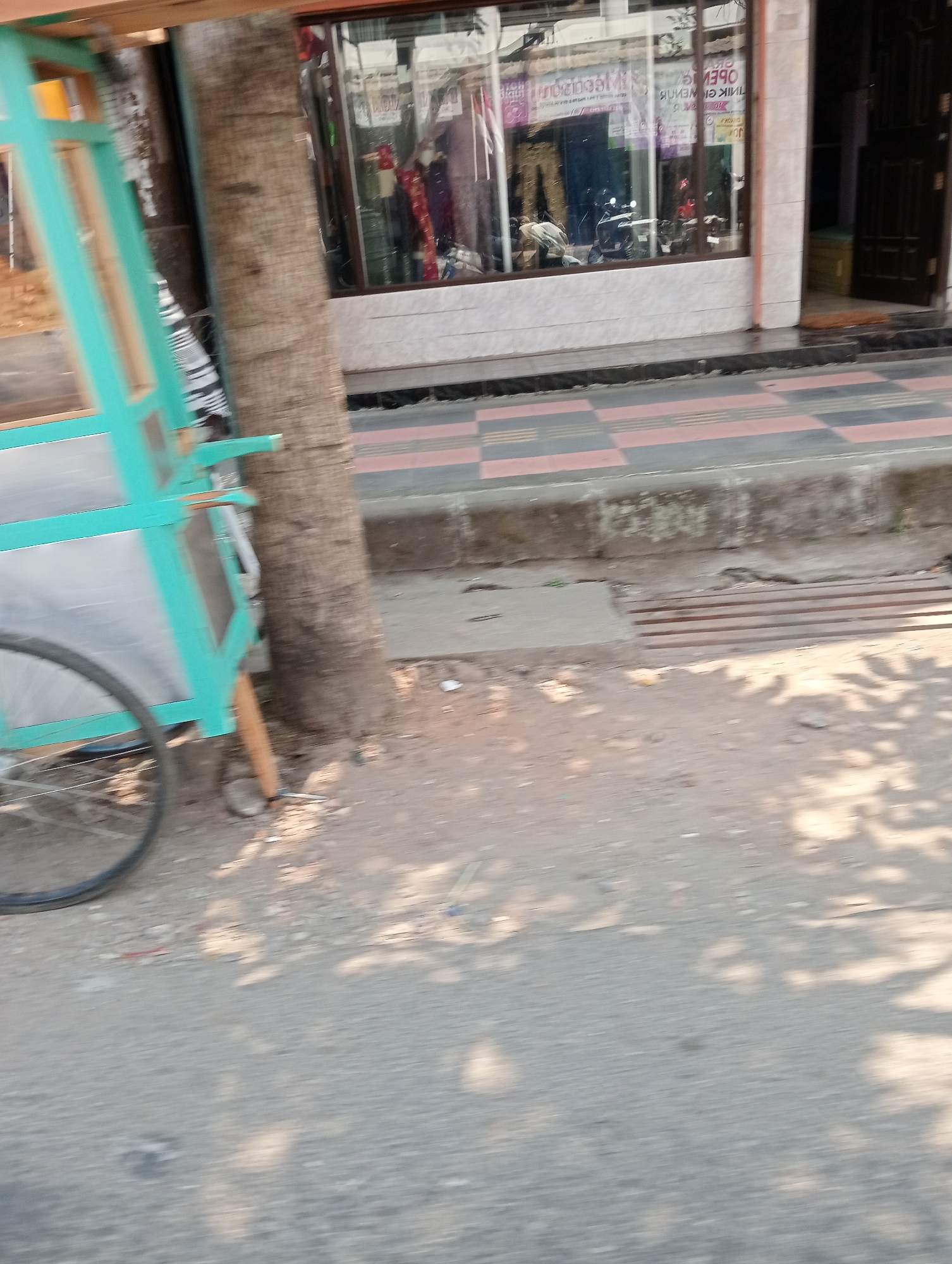 A street scene in Grabag, Indonesia, shows a turquoise cart with a bicycle wheel on its left side, positioned next to a tree trunk. Behind the cart and tree, a storefront with a large glass window displays various clothing items. The storefront is tiled with white and pink squares on the lower portion of the facade, and above the tiles, the window is framed by dark trim. The ground in front of the storefront is paved with a pattern of pink and black squares. To the right of the storefront is a dark wooden door. The street itself is made of asphalt, and dappled sunlight from overhead foliage creates shadows on the ground.