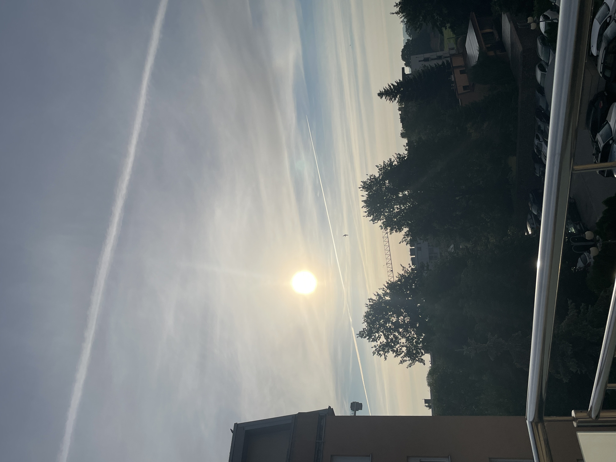 The image shows a clear sky with two contrails from airplanes. The sun is visible and bright, creating a lens flare. In the foreground, there is a balcony railing. Behind the railing, trees and buildings are visible. A parking lot with several cars is also present. A crane is visible in the distance.