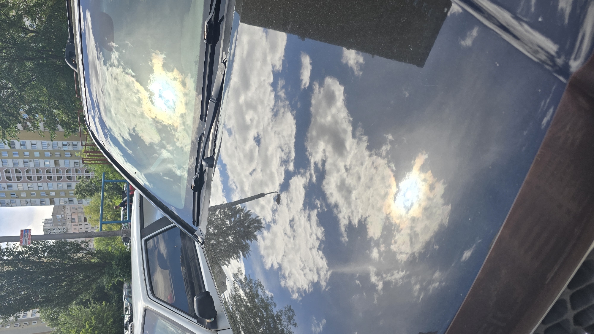 The image captures the front section of a dark-colored car, primarily focusing on its windshield and hood, which act as a highly reflective surface. The dominant feature is the vivid reflection of a bright blue sky filled with scattered white clouds and the intensely shining sun. The sun's reflection appears as a bright, slightly rainbow-hued spot.
In the background, tall, multi-story apartment buildings with numerous windows line the left side of the frame (when oriented correctly). Lush green trees are visible between the car and the buildings, and a street light with its pole is also reflected on the car's hood and visible in the background. A blue metal structure, possibly part of a fence or playground, is also present between the trees and the buildings.
The scene takes place on a clear, sunny day, indicated by the strong sunlight and well-defined cloud formations reflected on the car. No people are visible, and no specific activity is occurring; the image depicts a static street scene.
A rectangular sign, partially obscured by foliage in the mid-ground, displays visible text in Cyrillic characters. The word "ТАРІФИ" (Tariffs) is clearly discernible, followed by the numbers "10-90," suggesting an informational or parking rate sign. The overall setting appears to be an urban or suburban residential area.