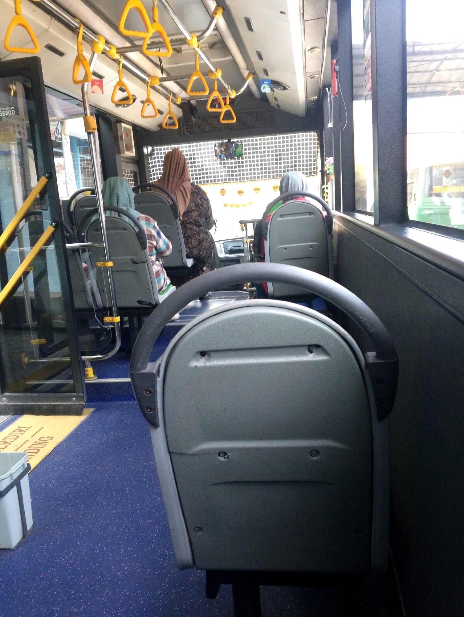 An indoor view of a bus interior shows multiple seats and passengers. Yellow handrails are attached to the ceiling, and there are also grey and black handrails on the seats. Several passengers are visible, mostly wearing headscarves. The floor is blue with small flecks of color. A yellow mat with text is on the floor near the entrance. The windows show an outdoor scene that is out of focus.