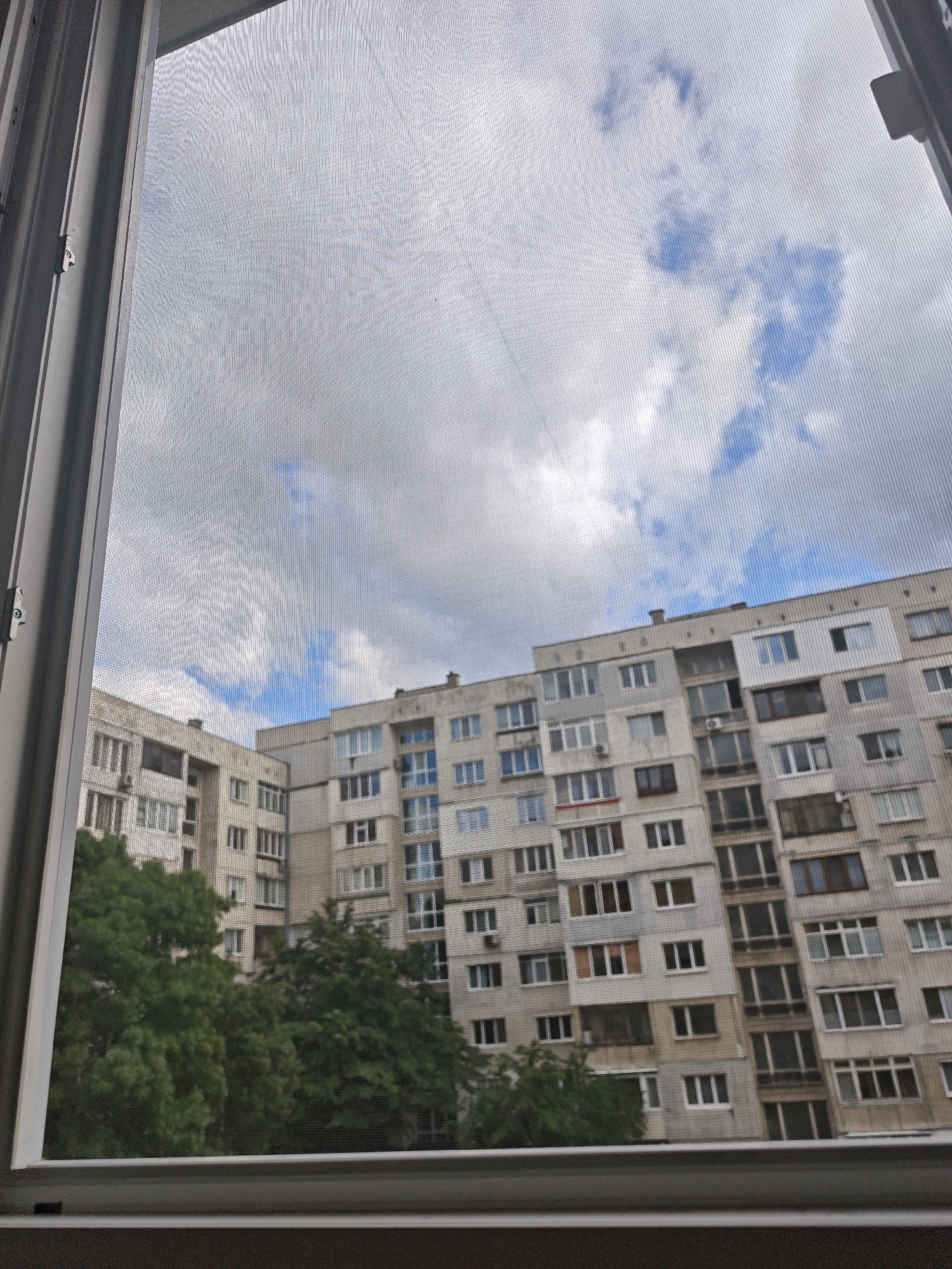The image presents a view from inside a building, looking out through a window in an urban residential area of Sofia, Bulgaria. The entire outdoor scene is observed through a fine, grey mesh window screen, which overlays a distinct grid pattern onto the view. A light-colored window frame is visible on the left and bottom edges, framing the scene.
The main subjects are two large, multi-story apartment buildings. Their facades are a mix of light grey and white, characterized by numerous windows of varying designs and sizes, some featuring balconies or enclosed glass areas. Several air conditioning units are affixed to the exterior walls. The architectural style is typical of Eastern European residential blocks.
Below the buildings, the upper canopies of vibrant green trees are visible, providing a natural element within the urban setting. The sky above is partly cloudy, with substantial patches of blue sky peeking through white and grey clouds, indicating it is daytime. There is no visible activity or people in the scene, which appears calm and static. No text is visible.