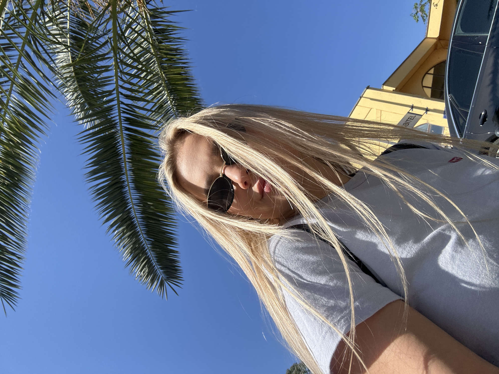 The image presents a low-angle, tilted perspective of a young person with long, flowing blonde hair. Her face is angled downwards towards the camera, with dark sunglasses obscuring her eyes. Her lips are slightly parted, and she wears a light-colored short-sleeved t-shirt, featuring a small red logo on the left chest. Bright sunlight creates strong highlights on her hair and illuminates the scene.
The background is dominated by a clear, vivid blue sky. Prominent large green palm fronds fill the upper left and extend across the top, indicating a tropical or Mediterranean environment. To the upper right, a light yellow or beige building with an arched window and dark trim is visible. A dark-colored vehicle, likely a car, is partially visible on the far right, adjacent to the building. Partial text "BITEL" is discernible on a sign attached to the building.
The scene takes place outdoors during bright daylight, likely midday or early afternoon, evidenced by the strong sun and defined shadows on the person's t-shirt. The weather is clear and sunny. The warm, sunny outdoor setting is consistent with a Mediterranean location such as Budva, Montenegro. The tilted camera angle suggests a dynamic selfie or a low-angle candid shot.