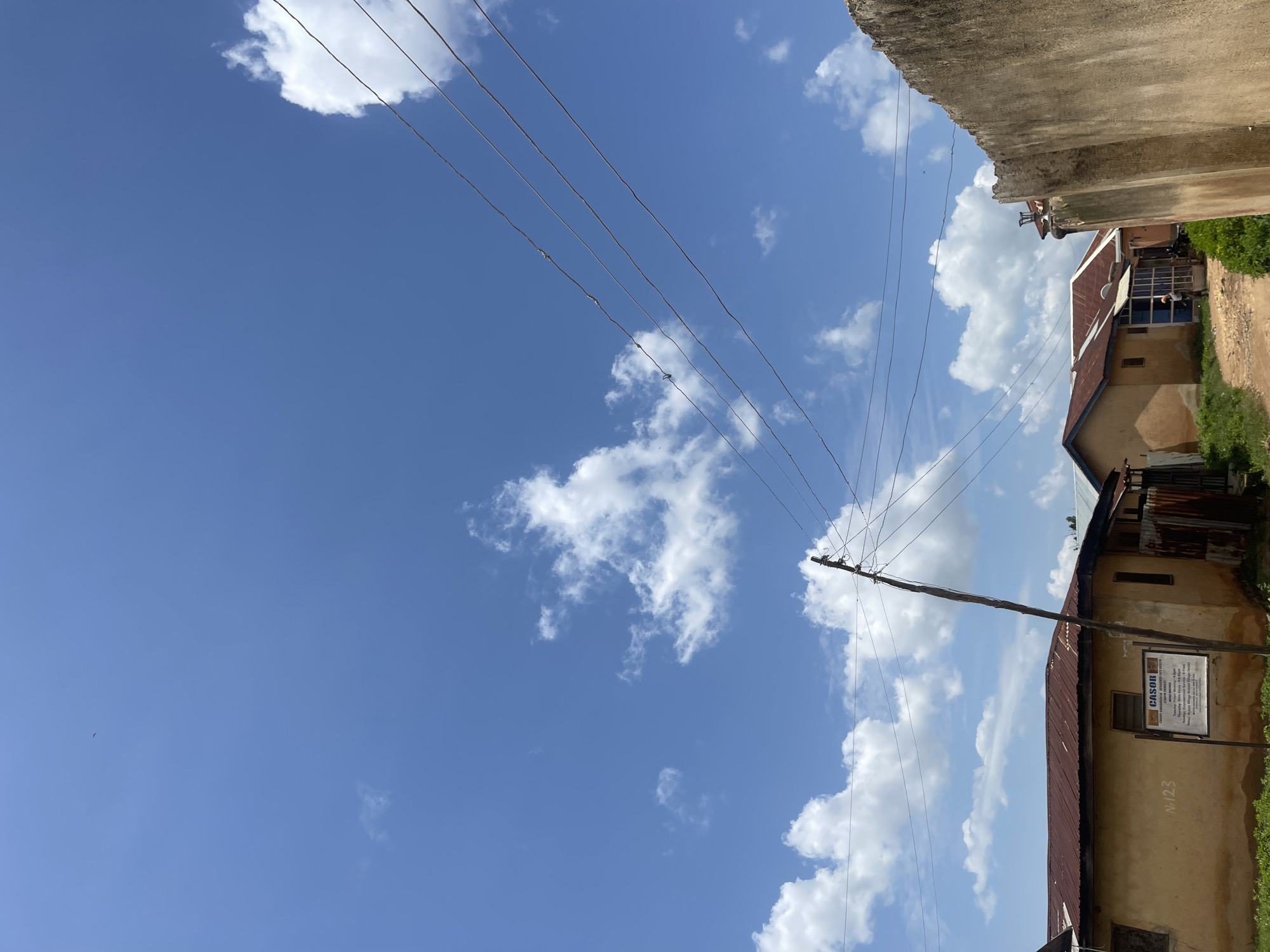 This is a ground-level, outdoor, daytime shot of a rural or semi-rural setting, likely in the city of Rafin Par, Nigeria, under a bright blue sky with scattered white clouds. The image is dominated by overhead electrical wires stretching diagonally across the frame, supported by a single wooden pole.
In the foreground and midground, several single-story buildings with corrugated metal roofs are visible. The walls of these buildings appear to be made of earth or stucco, painted in muted yellow and beige tones. One building, on the left, has a sign with text on it, including the word "CASOR," and the number "N.123". There is also a corrugated metal fence or gate. A dirt path winds between the buildings. Green vegetation, including grass and some bushes, is present around the buildings and along the path. A tall, rough-textured concrete wall occupies the far right of the frame.
In the background, the sky is clear and blue, with fluffy white cumulus clouds. The overall impression is one of a sunny day in a modest, possibly developing, community.
