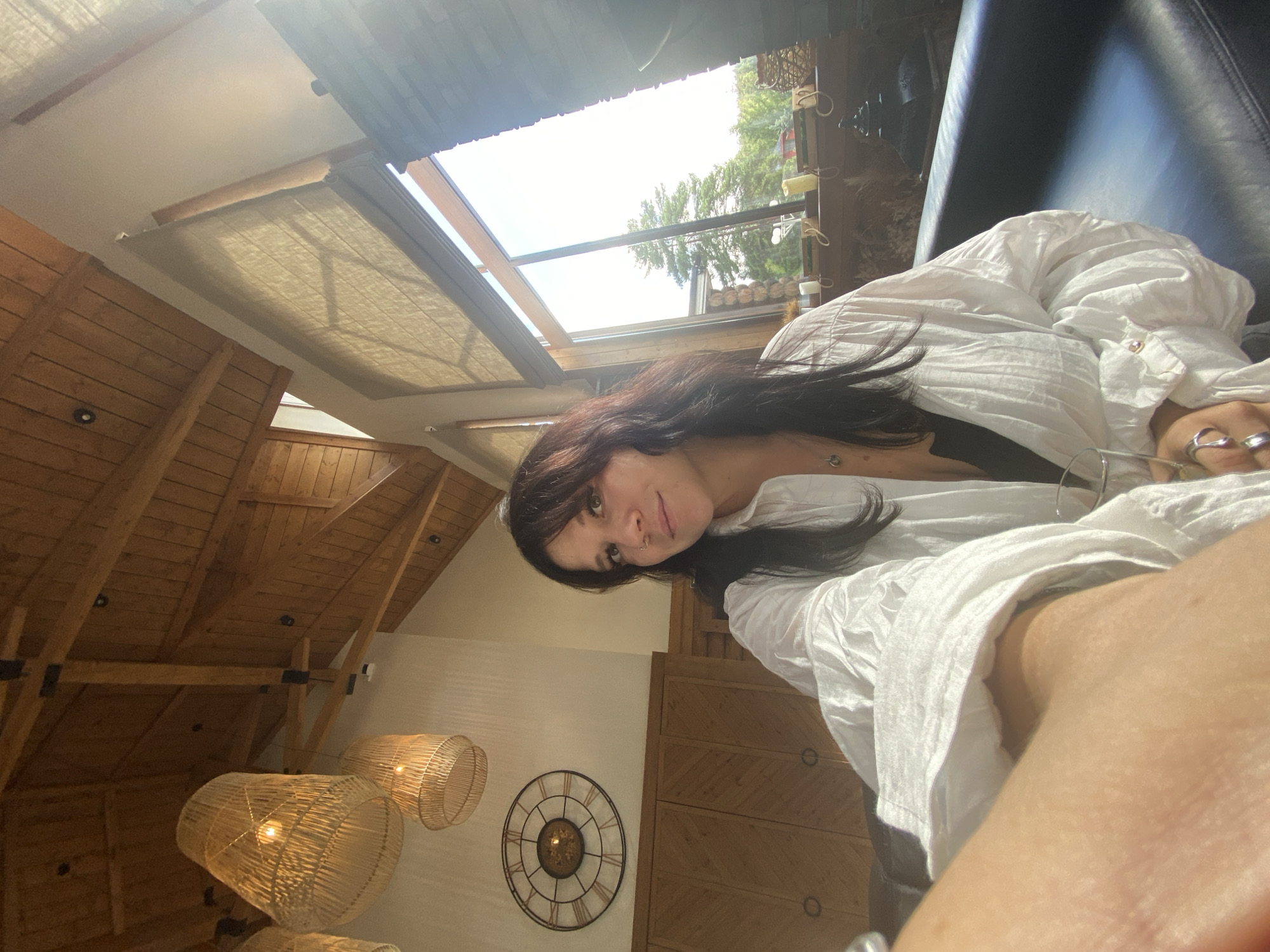 A woman with long, dark hair and bangs poses for a selfie in a room with a high, wooden-beamed ceiling. She wears a white, oversized shirt with billowy sleeves and a black top underneath. A pendant necklace is visible. The room features large windows that let in bright sunlight, a stone fireplace, and wicker lamps hanging from the ceiling. A large, round clock with Roman numerals hangs on the wall above wooden cabinets. The woman is seated on a dark leather couch. The background visible through the windows shows trees and a rustic building. The environment suggests a spacious, possibly rural or vacation-style home.