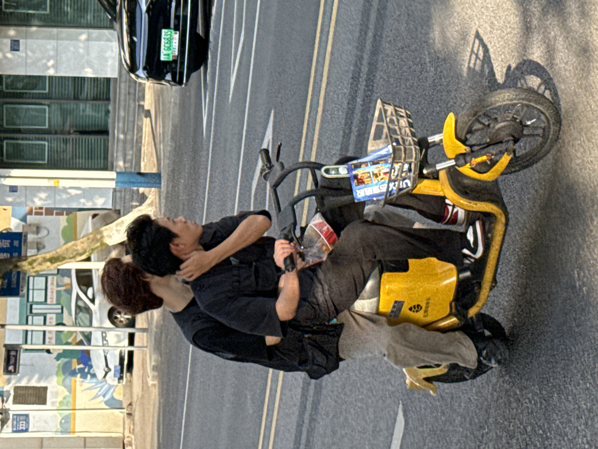 The image depicts two individuals riding a bright yellow electric scooter on an urban street in what is indicated to be Jiangjin, China. The scene is captured from a side perspective, showing the scooter and its occupants in motion.
The subjects are two men. The man in front is driving the scooter, wearing a dark long-sleeved shirt and dark pants. The man riding pillion behind him has his right arm wrapped around the driver's waist. He is also dressed in dark attire, including a dark jacket or shirt and light-colored pants. Their expressions appear neutral as they navigate the street.
The primary object is a distinctive yellow electric scooter, possibly a shared mobility service vehicle, given the front basket and visible branding. The word "SELECT" and what appears to be a partial Chinese character (possibly from a brand like Hellobike) are visible on the scooter's body. The front basket contains various items and possibly advertisements. In the background, on an adjacent lane, a black car is visible. Its license plate, "沪A GCB635," identifies it as registered in Shanghai.
The setting is a well-maintained asphalt road featuring distinct white and yellow lane markings. Modern multi-story buildings with numerous windows line the left side of the street. Some greenery, including parts of a tree trunk and branches, is also visible. The lighting suggests it is daytime, with clear skies, indicated by the strong sunlight and visible shadows cast by the scooter and its front wheel.