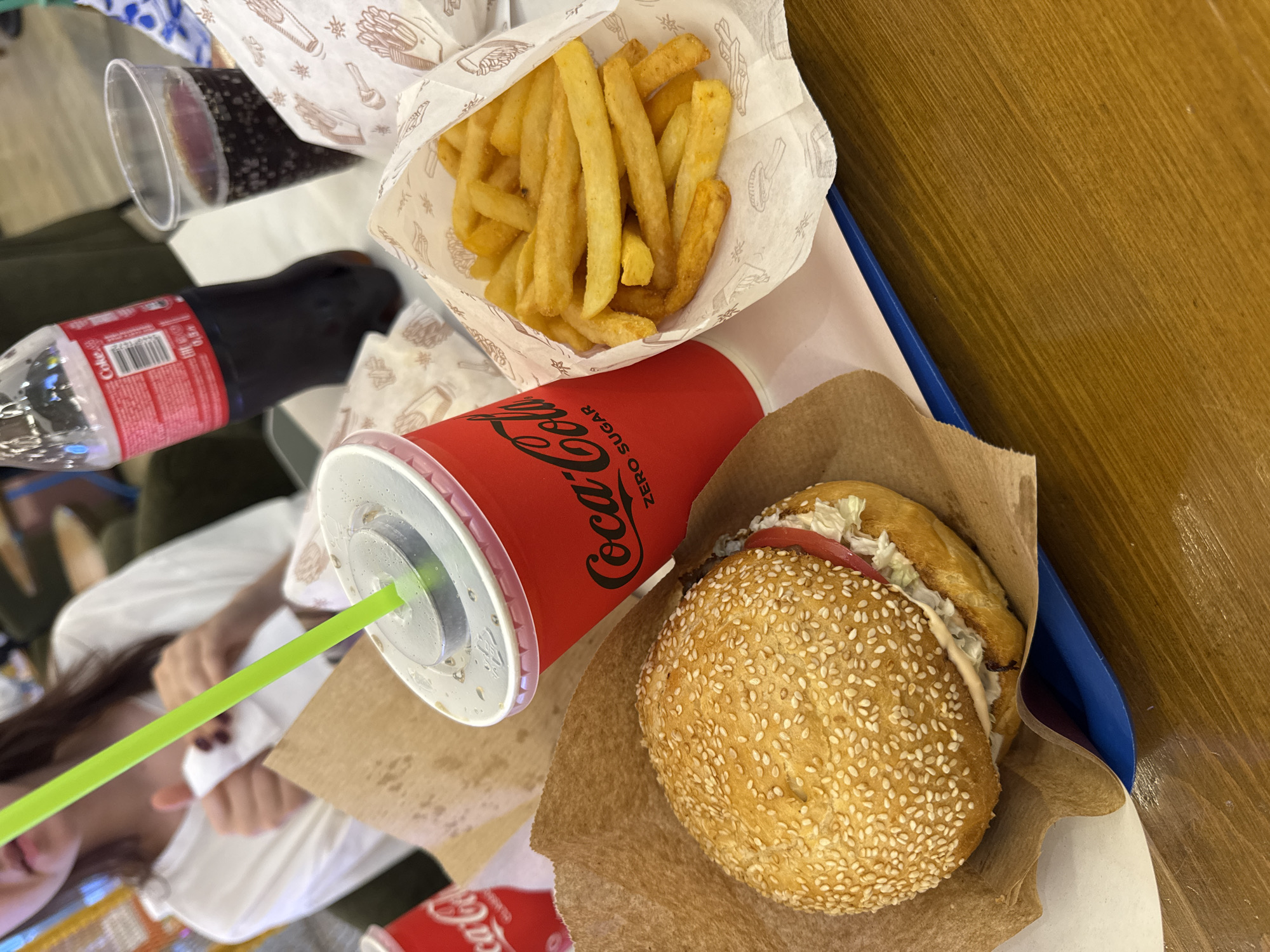In Oskemen, Kazakhstan, a table at a fast-food establishment is set with a meal. The meal includes a burger served in a brown paper wrapper, a cup of Coca-Cola Zero Sugar with a green straw, a separate paper container filled with French fries, and a plastic cup with a dark beverage. A partially visible person is in the background, wearing a white t-shirt and holding a white napkin. The table surface is made of wood.