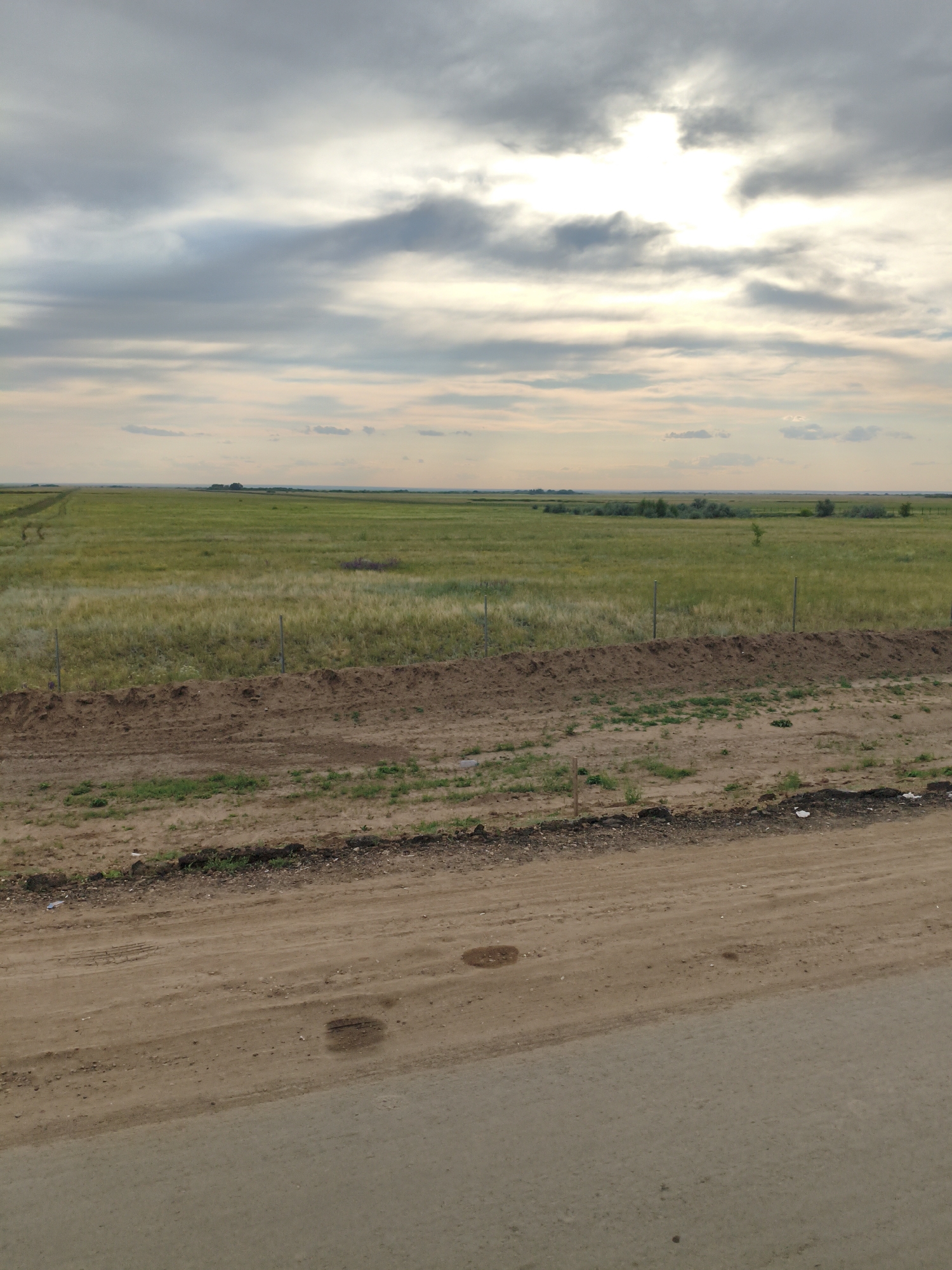 The image depicts a vast, open grassland under a cloudy sky. The foreground is a dusty dirt road with some patchy vegetation. Behind the road, there is a raised strip of soil, possibly a berm, with a simple wire fence running along its crest. Beyond the fence, a large expanse of green and yellowing grass stretches to the horizon, punctuated by distant trees and shrubs. The sky is overcast with patches of bright sunlight breaking through the clouds, suggesting either dawn or dusk. The scene is devoid of people or any significant structures, presenting a natural, undeveloped landscape. Based on the metadata, this image was taken in Teploe, Russia.
