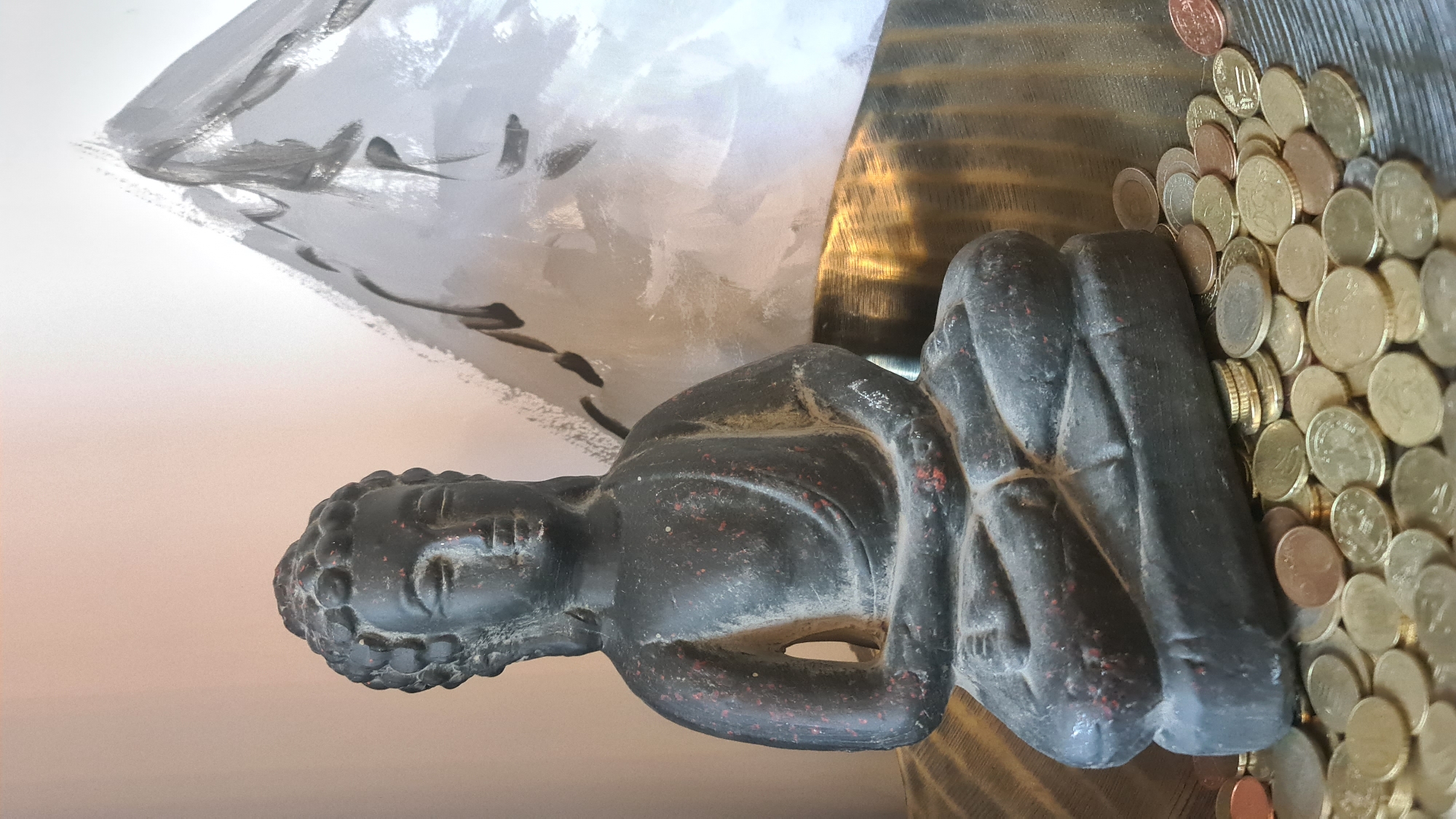A close-up shot shows a small, dark-colored statue of Buddha in a seated, meditative pose. The statue has a worn, textured surface with hints of red detailing. To the right of the statue, there's a cluster of Euro coins of various denominations. The background includes a shiny, gold-colored, textured surface and an abstract painting in shades of gray and white. The light in the scene is diffused, casting soft shadows. The location is unknown.