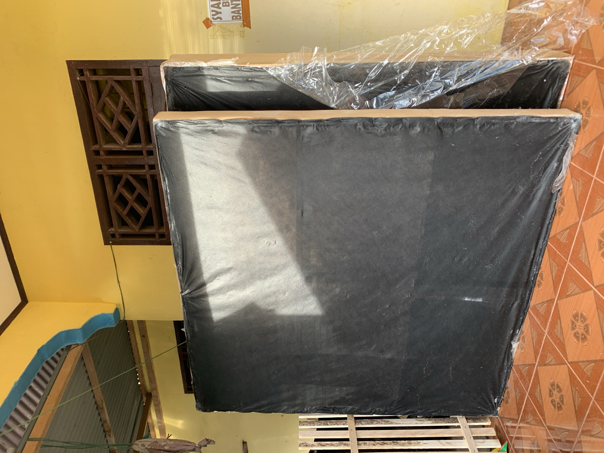 Two large, black upholstered items, possibly mattresses or large cushions, are leaning against a yellow wall. The items are wrapped in plastic sheeting, with one item more securely covered than the other. The foreground is dominated by a patterned orange tile floor. To the left of the upholstered items is a section of light-colored wooden fencing. Above and behind the items, part of a corrugated metal roof is visible, supported by wooden beams. A decorative wooden lattice is set into the yellow wall. On the wall to the right, a piece of orange tape with partial text in what appears to be Indonesian is affixed. The scene suggests an outdoor or semi-outdoor storage area, possibly a home's exterior wall or patio in Meunasahdrang, Indonesia.