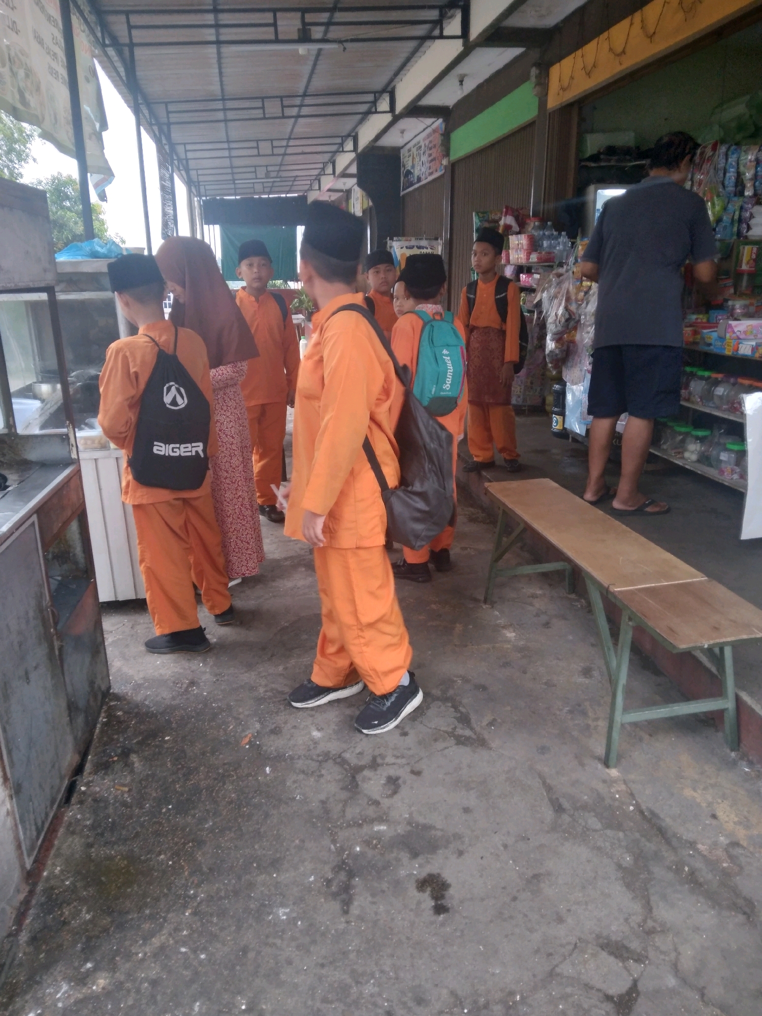 The image depicts a group of children, likely students, wearing orange uniforms and black caps, standing in front of a shop in Batam, Indonesia. A woman in a patterned dress and headscarf is also present. One boy has a black drawstring bag with the brand "aiger" printed on it, and another has a teal backpack. A man in a dark grey t-shirt and dark shorts is browsing items inside the shop, which appears to be a convenience store or small market selling packaged goods and drinks. A wooden bench is positioned in front of the shop. The environment suggests an outdoor covered area, possibly a walkway or storefront. The ground is concrete and appears somewhat worn.