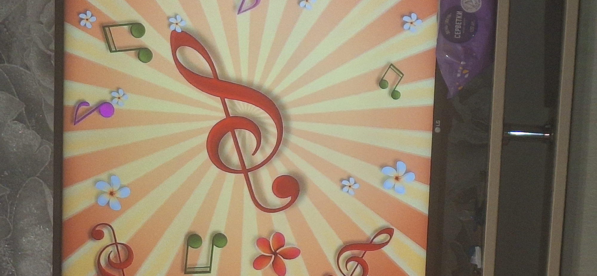 The image depicts a close-up of a television screen displaying a colorful, whimsical graphic. The graphic features a large red treble clef as the central element, surrounded by stylized musical notes in green and purple, as well as light blue and pink flowers. The background is a radial pattern of yellow and orange stripes. The bottom of the screen shows the LG logo and a purple package with Russian text that appears to be "СЕРБЕТКИ," which translates to "wipes." The television is placed on a shelf in what appears to be an indoor setting, with a textured surface visible above the screen. There are no people or specific locations in Bila Tserkva, Ukraine identifiable in this image.