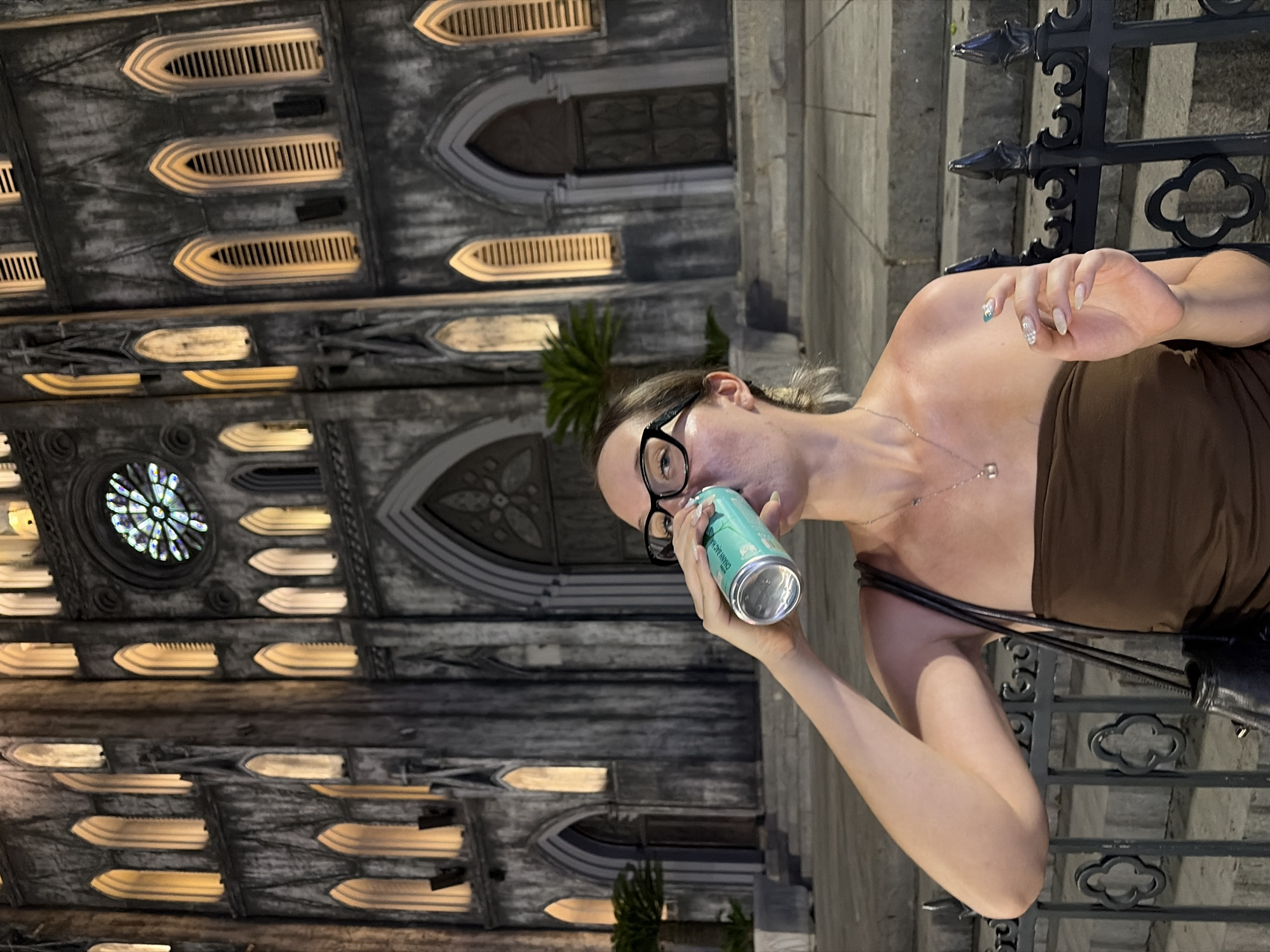 A young woman with blonde hair and glasses is drinking from a light blue can in front of St. Joseph's Cathedral in Hanoi, Vietnam. The cathedral, a neo-Gothic Roman Catholic building, is illuminated at night, with warm lights highlighting its arched windows and intricate stonework. The woman is wearing a brown strapless top and a silver necklace. She is standing near a black metal fence, with steps leading up to the cathedral behind her. The scene suggests a casual evening outing in a historically significant urban setting.