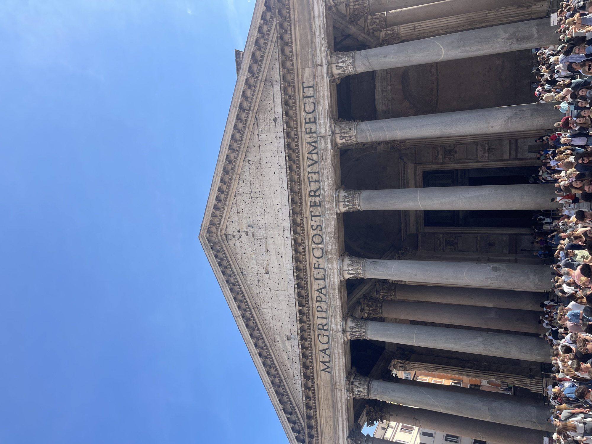 The image captures the Pantheon in Rome, Italy, from a low-angle perspective looking upwards. The imposing facade of the ancient Roman temple is prominently featured, showcasing its monumental columns, triangular pediment, and Latin inscription. The inscription reads "M·AGRIPPA·L·F·COS·TERTIVM·FECIT," indicating that it was built by Marcus Agrippa. A large crowd of people is gathered near the entrance, suggesting a busy tourist destination. The sky is a clear blue. The architectural details of the Pantheon appear weathered but well-preserved, indicating its historical significance.