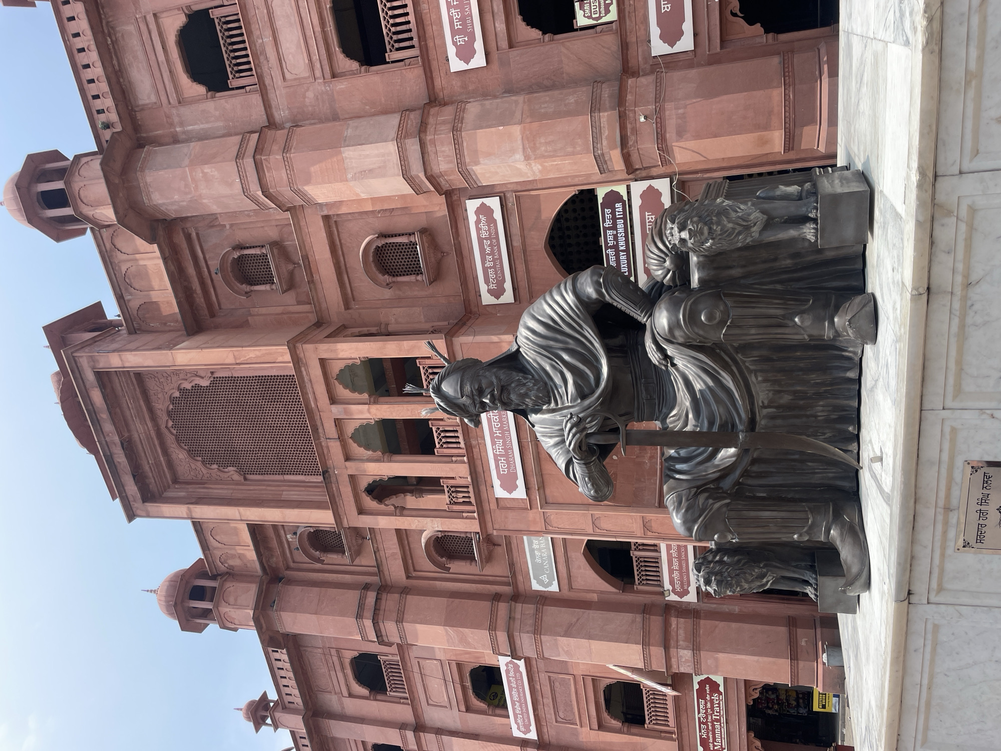 The image shows a large bronze statue of a historical figure seated on a throne, with two lion statues flanking the throne. The statue is positioned in front of a large, ornate building constructed with red sandstone. The building features intricate architectural details, including arched windows and decorative carvings. Several signs are visible on the building's facade, some in a script that appears to be Punjabi and others in English, indicating various establishments such as a bank and an insurance company. The statue and building are situated outdoors under a clear, bright sky. The overall scene suggests a historical or cultural landmark in Amritsar, India, possibly located near a market or commercial area given the presence of business signs. The statue itself depicts a person dressed in traditional attire, with a turban, beard, and holding a sword, indicating a figure of significance.