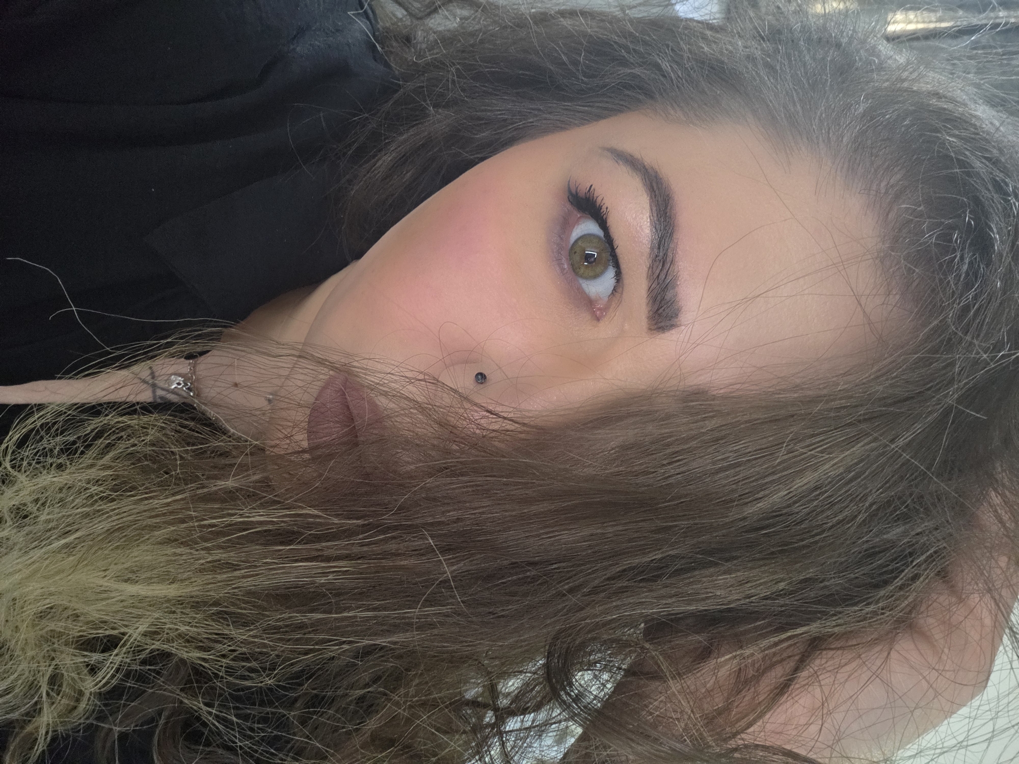 A close-up portrait of a woman lying down, her head angled towards the camera. Her right eye is prominent, showing an amber-colored iris. Her skin tone is fair with visible blush on her cheek. She has a piercing near the corner of her mouth. Her hair is a mix of brown and blonde, partially covering her face. She is wearing a black garment. The location is not discernible from the image.