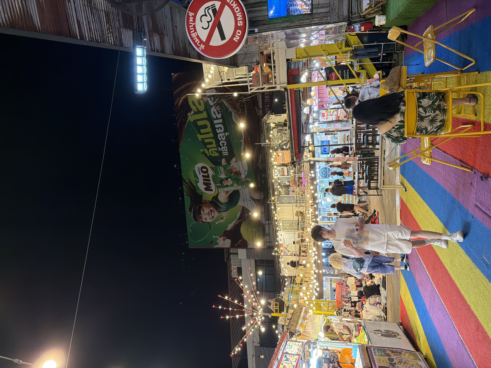 Captured at night, the image presents a bustling street scene in Phuket City Municipality, Thailand. Numerous stalls, adorned with strings of lights, line the pathway. Several people are walking around and dining. The ground is painted with a rainbow pattern. A sign warns “No Smoking”. Above the stalls hangs a large MILO advertisement.