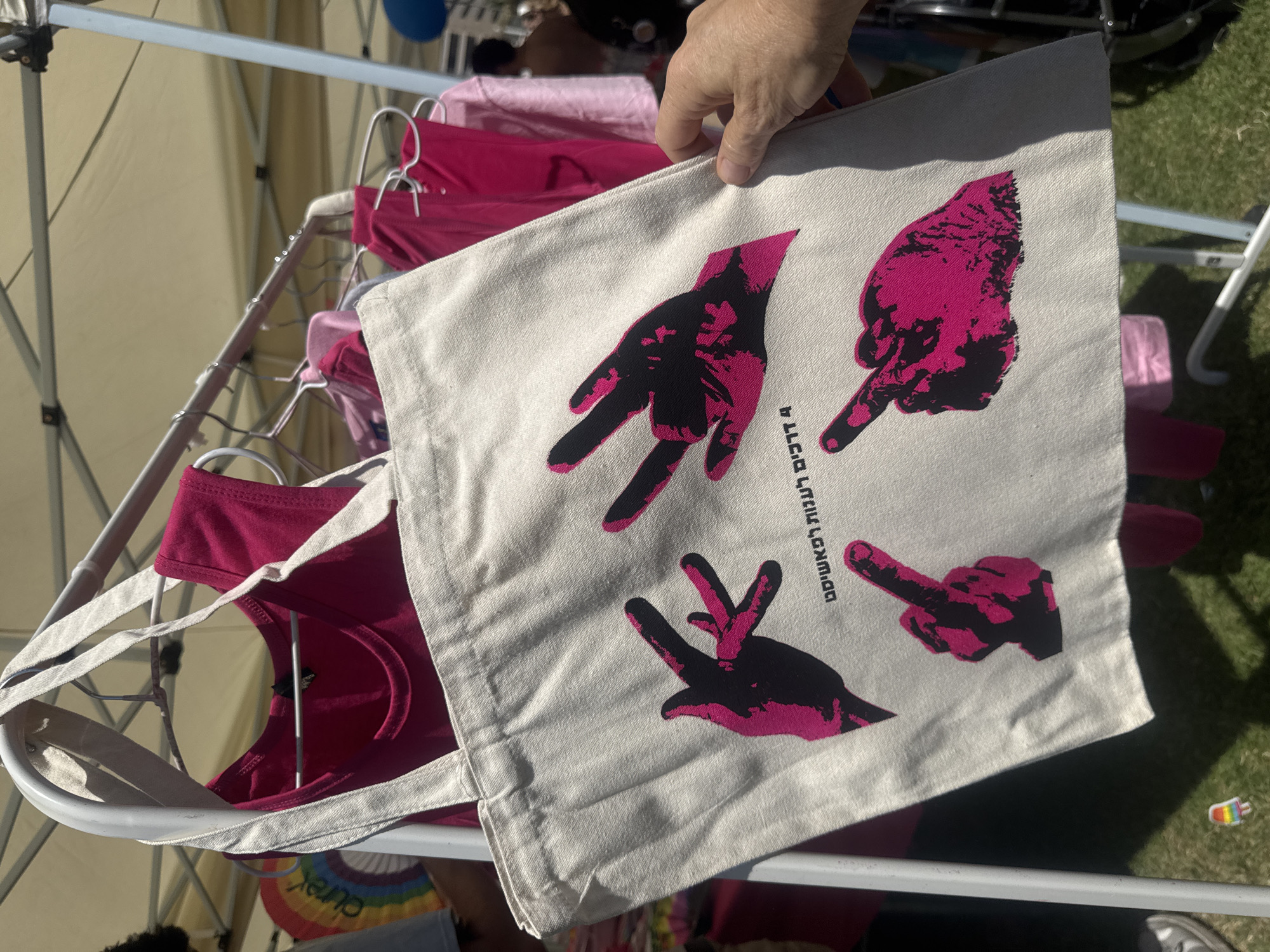 A canvas tote bag with a pink graphic of four hands making various gestures is displayed in the foreground. The hands are rendered in a stark, contrasting style of black and hot pink. Below the hands, text is printed in Hebrew, which reads "4 Ways to Dream." The bag is hanging on a white metal rack, with several pink garments, likely t-shirts or tank tops, also hanging on the same rack and behind it. The scene appears to be outdoors, possibly at a market or festival, with a blurred background showing tents and grass. A hand is visible on the right side of the image, holding the edge of the tote bag. In the lower left corner of the image, a small rainbow-colored popsicle-shaped sticker is visible on the ground. The location is identified as Tel Aviv, Israel.