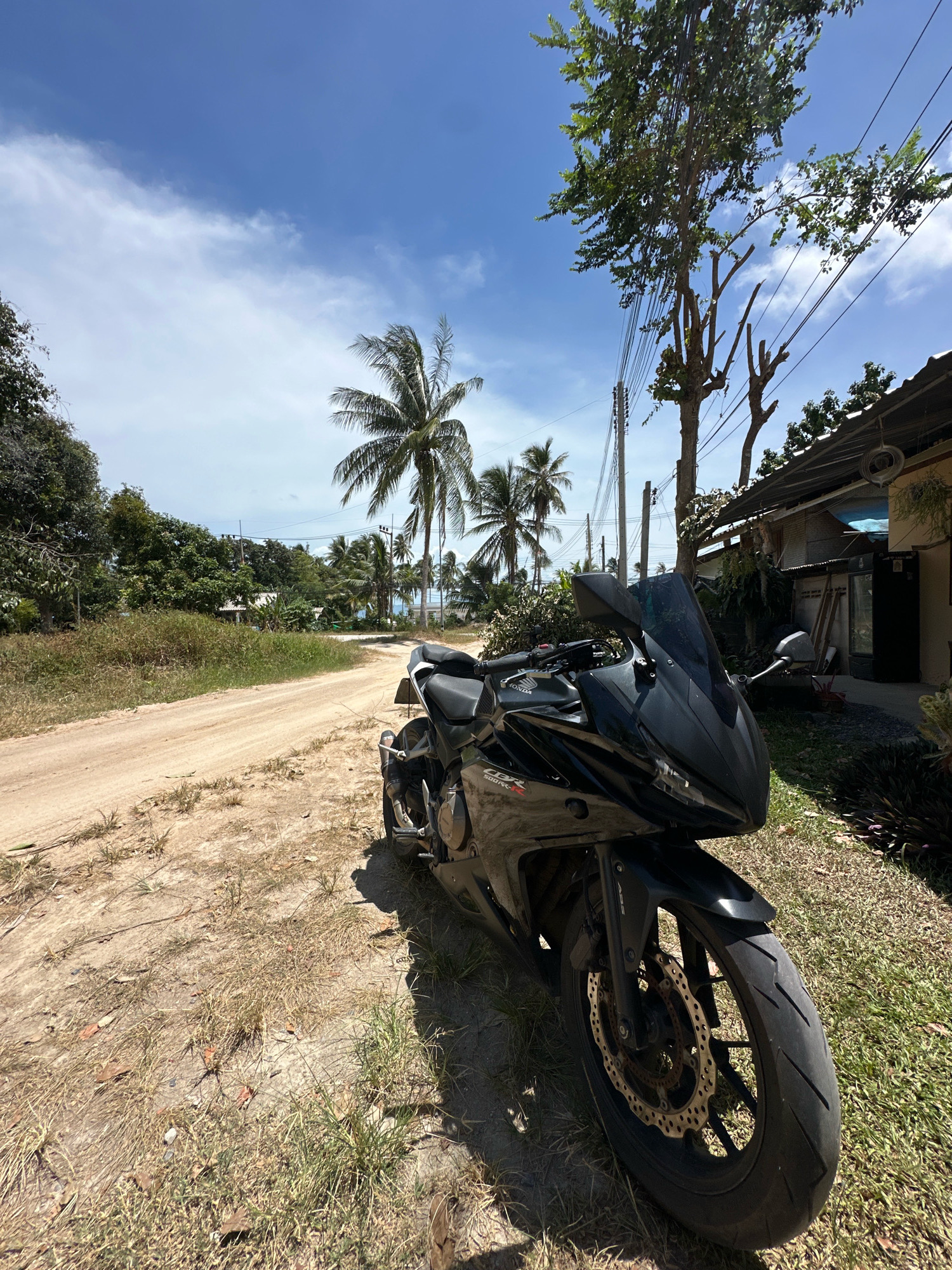 A black sport motorcycle is parked on the side of a dirt road in a tropical environment. The motorcycle is facing the viewer and is positioned on the right side of the image. The road is unpaved and appears to be in a rural or semi-rural area. In the background, there are palm trees, lush vegetation, and a building with a corrugated metal roof. Power lines are visible overhead, strung between poles. The sky is blue with some white clouds. The scene suggests a sunny day. The location is in the Ko Pha-ngan District of Thailand.