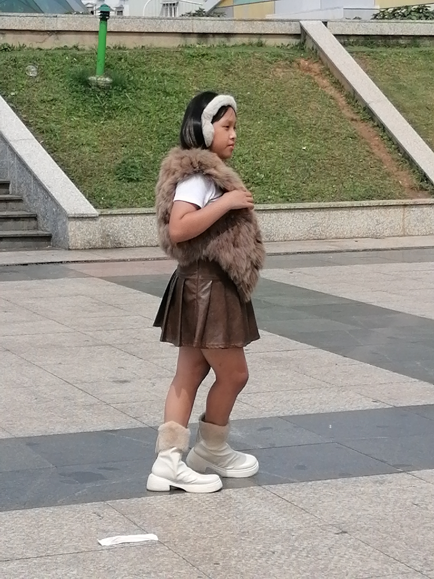 A young girl stands in a square paved with light gray and darker gray rectangular tiles. She is facing right, her body angled slightly away from the viewer. She has short, dark hair and is wearing a white t-shirt, a brown pleated skirt, and white boots with fur trim. She also has on a brown furry vest and a white furry headband. Behind her, a grassy slope rises, with a concrete retaining wall at its base. Stairs are visible to the left of the frame, ascending the slope. A green pole with a light fixture stands on the grass. The overall environment suggests an outdoor public space in Da Lat, Vietnam.