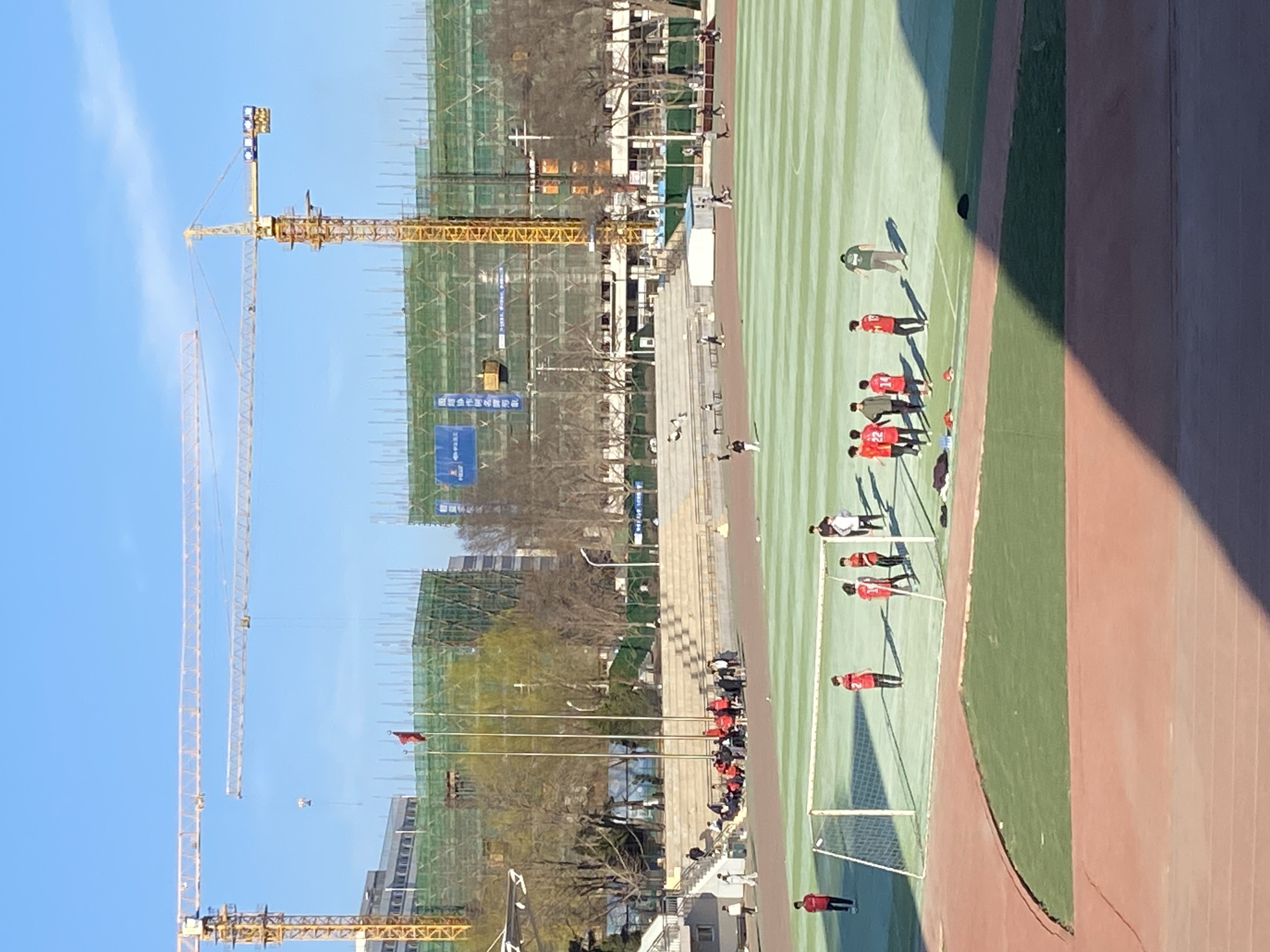 Here's a detailed description of the image:
The image depicts a construction site and athletic field scene, potentially located in or near the city of Zhongtun, China, given the presence of Chinese elements and common urban development features.
In the foreground is a portion of a soccer field with a group of people in red jerseys, likely a team training or playing. A soccer goal is visible. Alongside the soccer field, there is a running track.
In the background, there is a large construction site. Several cranes stand tall, indicating active building activity. Numerous buildings, either completed or under construction, are visible behind the construction equipment and scaffolding. Trees and other greenery are present, blending into the built environment. The sky is clear with a few clouds.