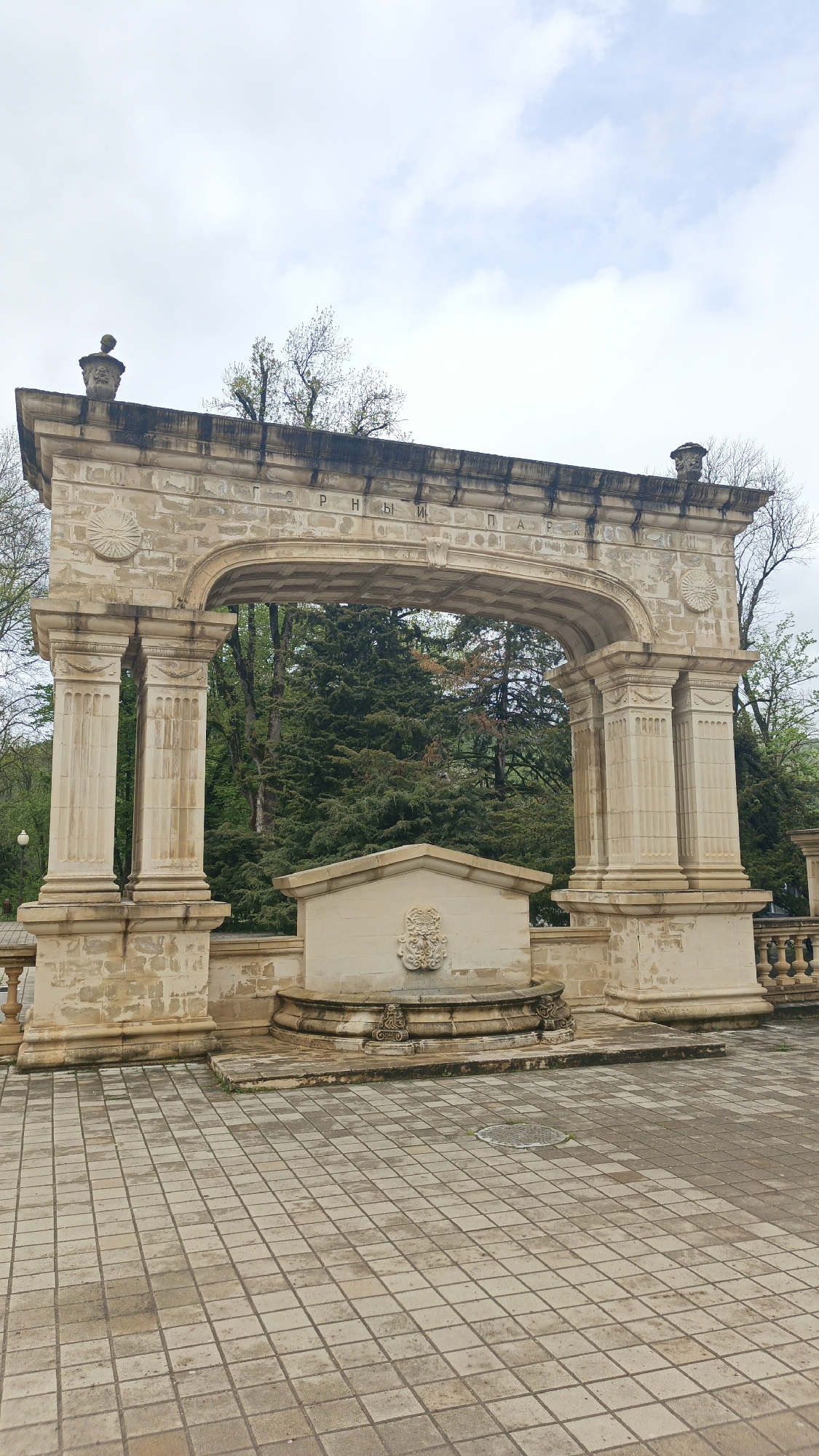 The image shows the Psekups park entrance arch in Goryachiy Klyuch, Russia. The arch is made of light-colored stone, supported by fluted columns, and topped with decorative elements. The inscription in Cyrillic letters on the arch reads "PARK". Behind the arch, green trees are visible. In the foreground, there's a stone fountain base and a tiled plaza. The sky is overcast. No people are visible.