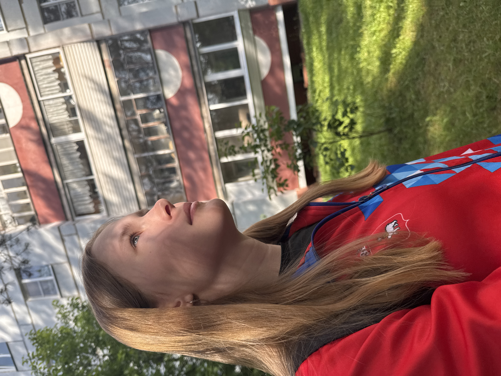 A young woman with long, light brown or blonde hair is depicted in a medium shot, looking upwards and to the left, in an outdoor setting in Chaikovsky, Russia. She is shown in profile, with a neutral and contemplative expression. She wears a bright red jacket with blue and white geometric patterns on the upper right arm/shoulder area, and a dark garment underneath. A small white logo, appearing to be a stylized bird or animal (possibly a hawk or eagle) within a red outline, is visible on the red jacket near her chest.
Behind the woman, to the left, stands a multi-story residential building with a light-colored facade interspersed with prominent vertical red sections. These red sections feature distinctive white circular architectural elements and numerous rectangular windows, some with white frames. To the right of the building and behind the woman, a vibrant green lawn or grassy area with trees and vegetation is visible, displaying elongated shadows.
The scene is illuminated by sunlight, suggesting either late afternoon or early morning, with warm light catching the woman's hair and face. The weather appears clear and sunny. The architecture of the building provides a location cue consistent with post-Soviet urban environments. No legible text is visible beyond the stylized logo on the jacket. The woman is not engaged in any specific activity beyond looking upwards, contributing to a calm, observational mood.