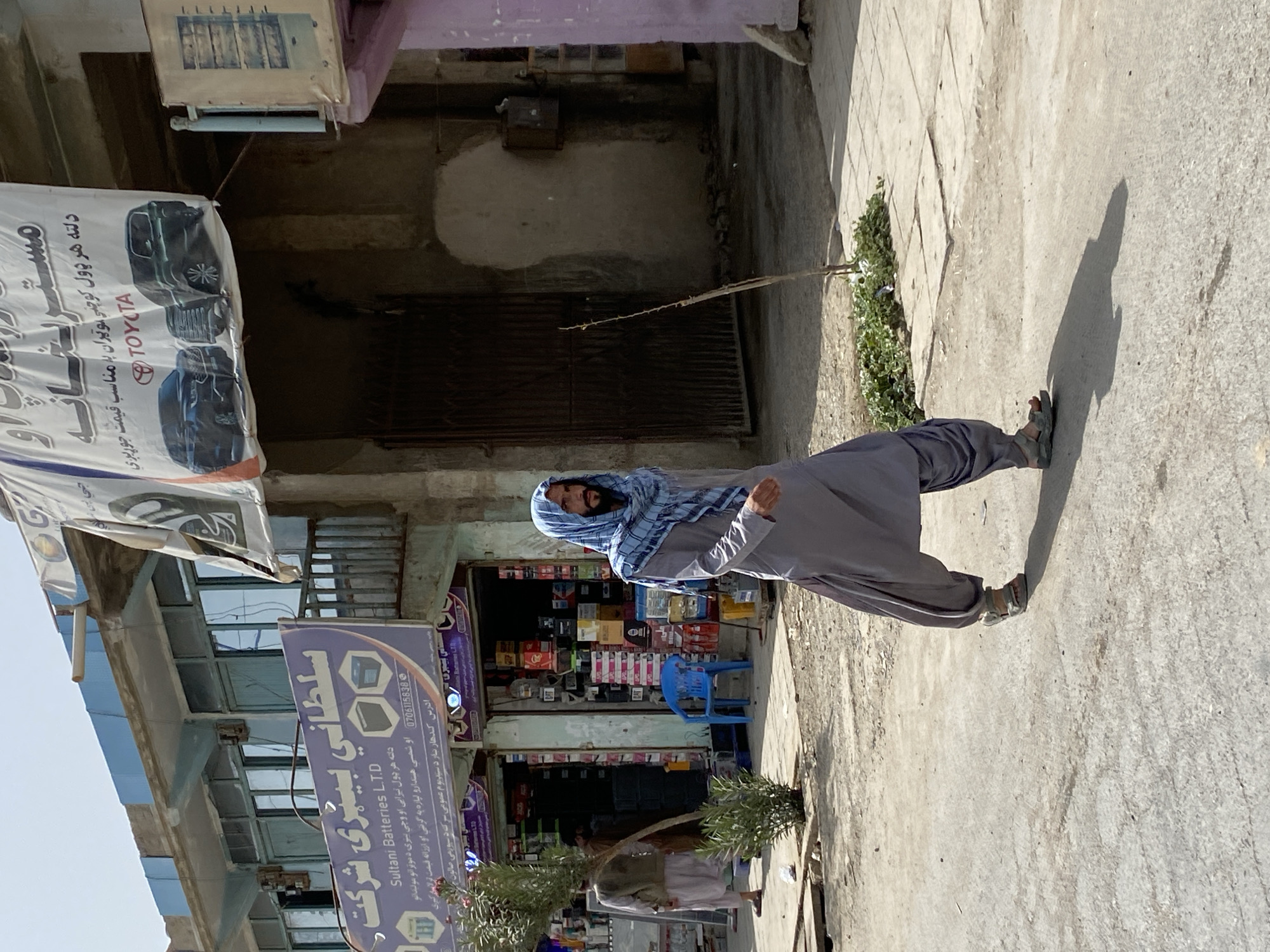 The image depicts a street scene in Kandahar, Afghanistan. In the foreground, a man wearing a grey shalwar kameez and a blue checkered scarf walks across a paved road. He has a beard and is wearing sandals. Behind him, a shop is visible with signage that appears to be in Arabic script. The shop displays numerous batteries and other items. To the left of the shop, another person is visible walking away from the camera. The background shows the facade of buildings with visible windows and some advertising banners. The overall environment appears to be an urban street with commercial activity.