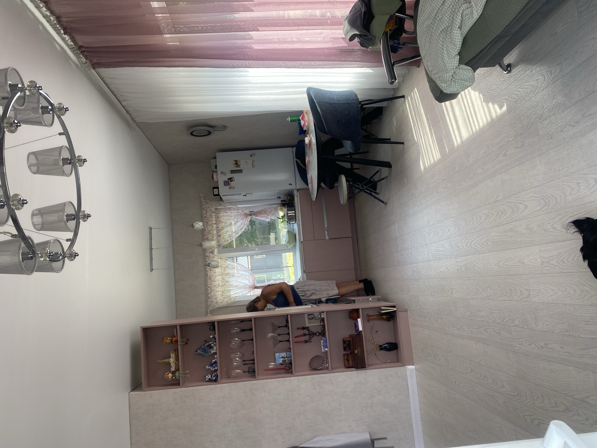 The image shows an interior scene that appears to be a kitchen and living area. The room has light-colored wooden flooring and light-colored walls, with some textured wallpaper on the left.
In the foreground on the right, a dark-colored sofa is partially visible, covered with a light-colored patterned blanket. Sunlight streams in from a window, creating distinct shadow patterns on the floor.
To the left, a tall, open shelving unit painted pink displays various decorative items, including vases, figurines, and glassware. A woman in a blue tank top and striped shorts is standing in front of the shelves, appearing to be organizing or looking at the items.
Further into the room, a kitchen area is visible. There are pink cabinets, a white refrigerator with items on its door, and a round table with two dark chairs. A small stool is placed near the table. Sheer pink and white curtains hang by a window, allowing natural light to enter. The overall impression is of a lived-in, domestic space. The mention of "Tret'ya Pad', Russia" is external information not discernible from the image itself.